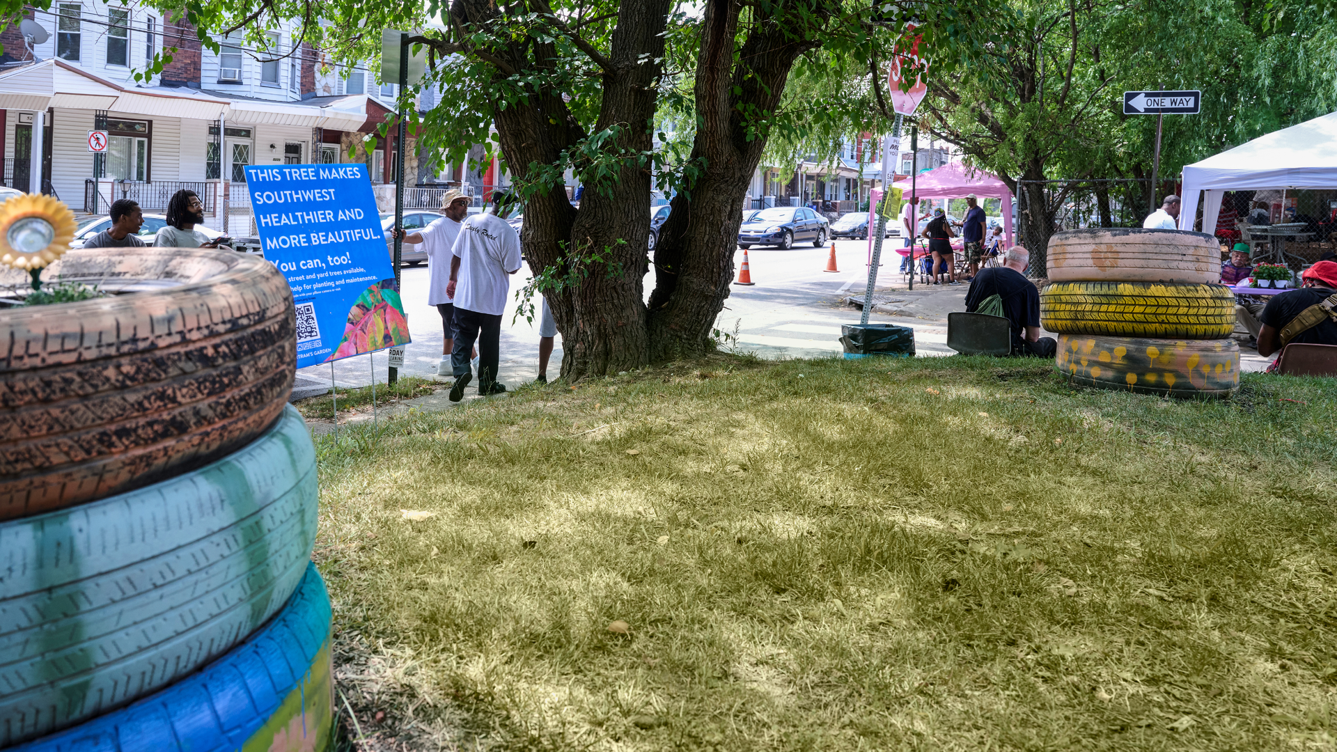 A grassy community garden on an urban corner includes a large sign next to a tree, “This tree makes Southwest more beautiful and you can too!”