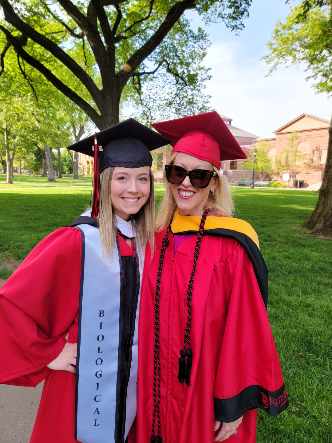 Regina Whittick and her daughter Charlotte, both smiling and wearing red graduation gowns on a college campus, Regina in a red graduation cap and Charlotte in a black cap