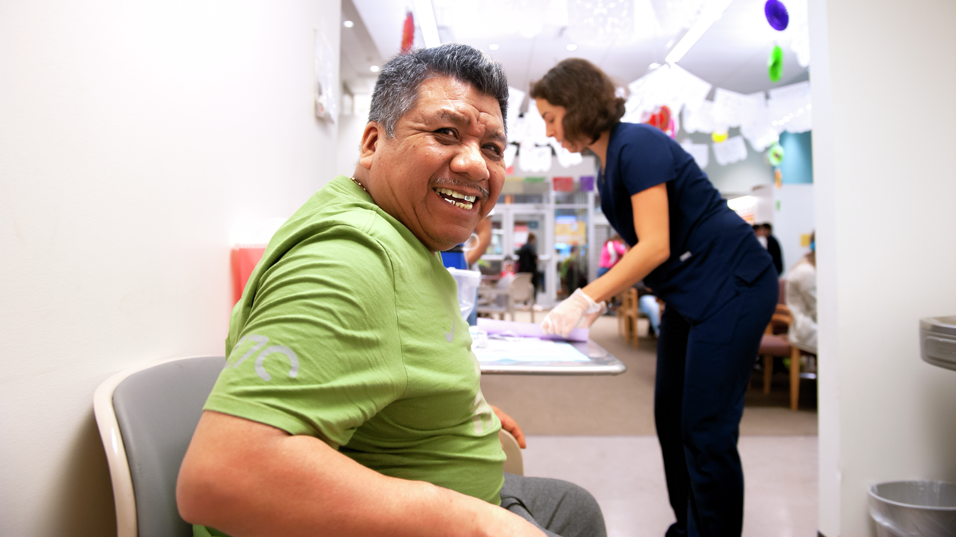 A seated middle-aged man smiles broadly while a nurse and the Puentes waiting room are in the background