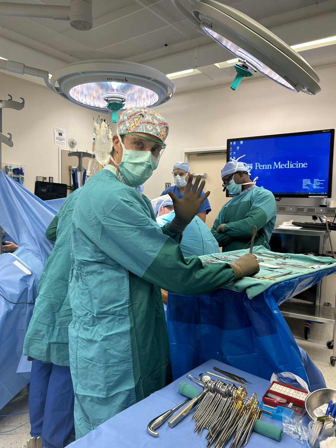 Chris Naimo, wearing a surgical gown, mask, and face shield, waves while standing next to an instrument tray in the OR