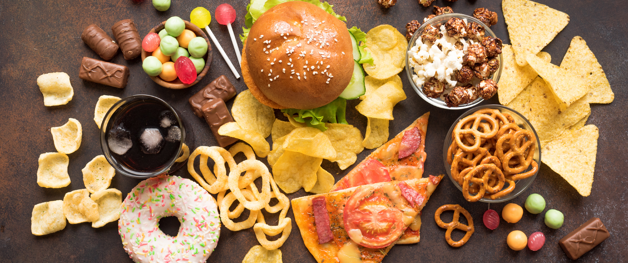 Food on a table, including a burger, pizza slices, chips, and candy