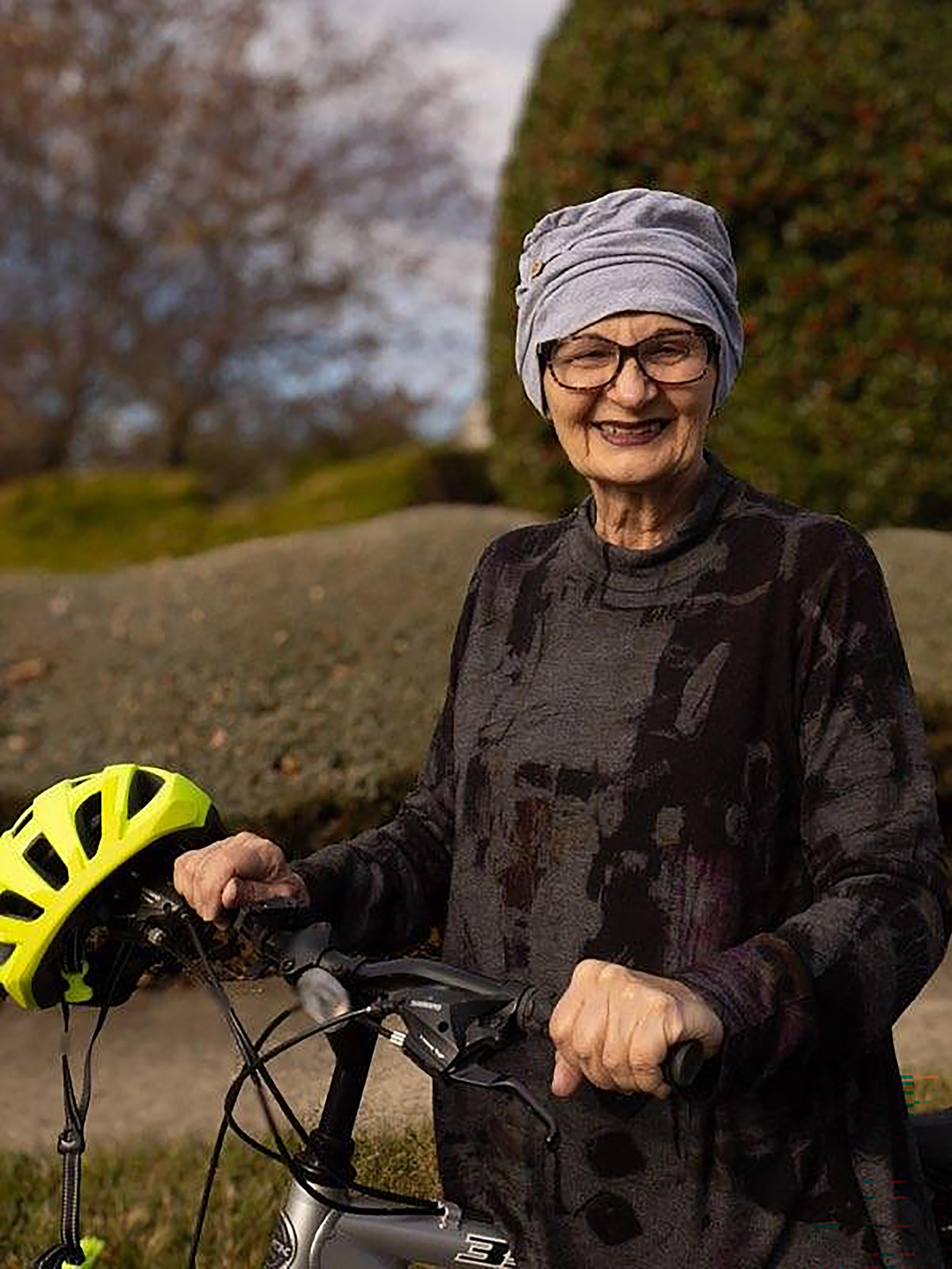 Donna Frandsen, an older woman wearing a  soft cap, stands beside a bicycle