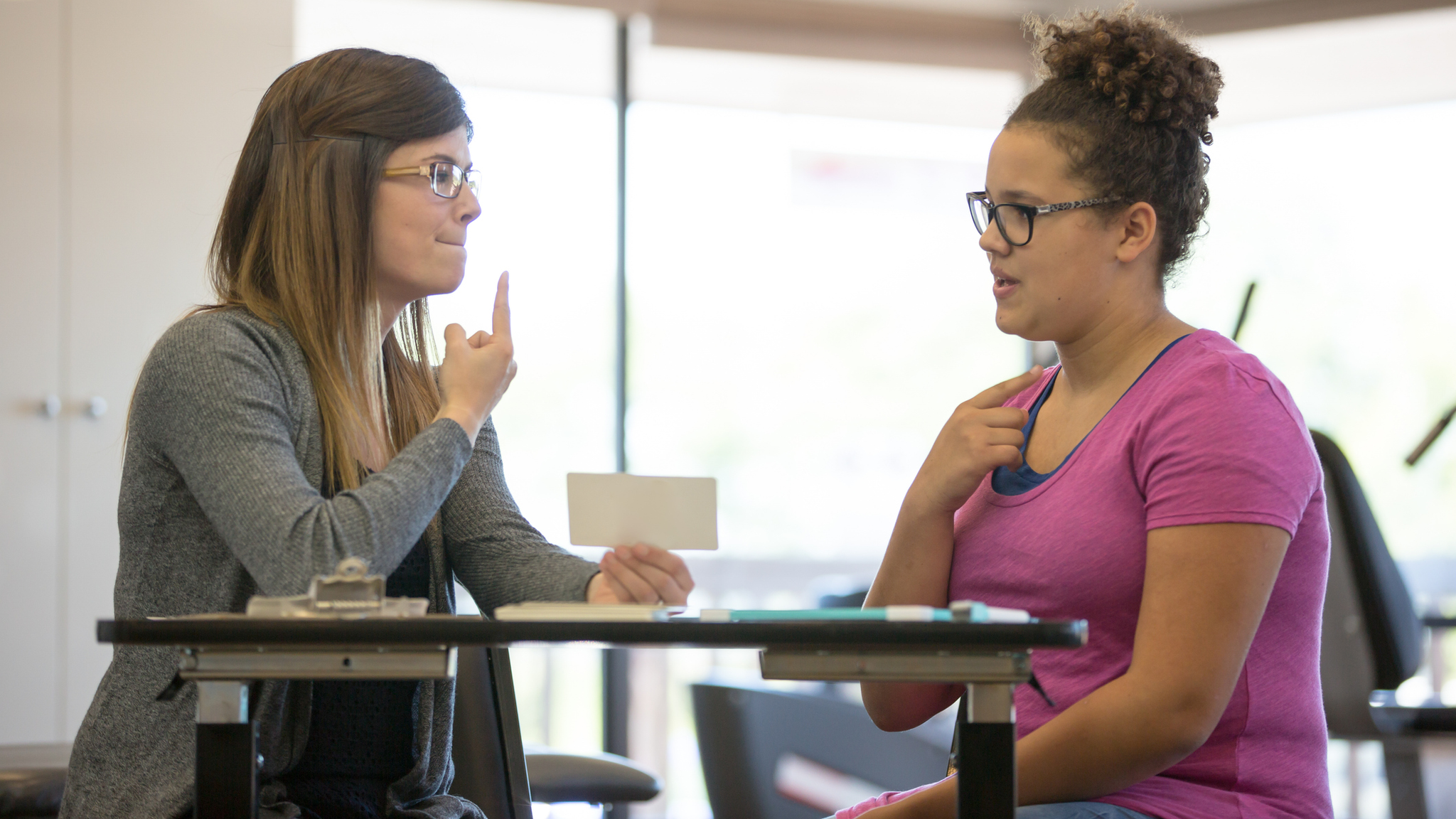 A young adult female speech therapist is helping a teenage patient inside of a clinic.