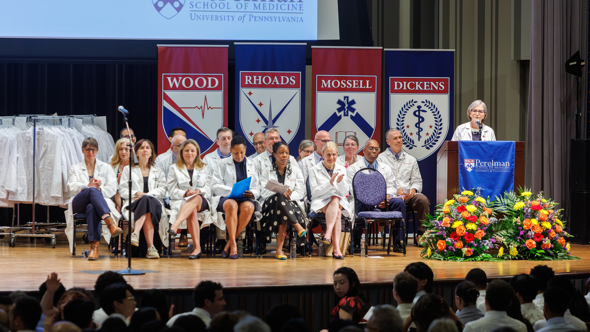Jennifer Kogan speaks at a podium while other medical school faculty sit on stage and listen 