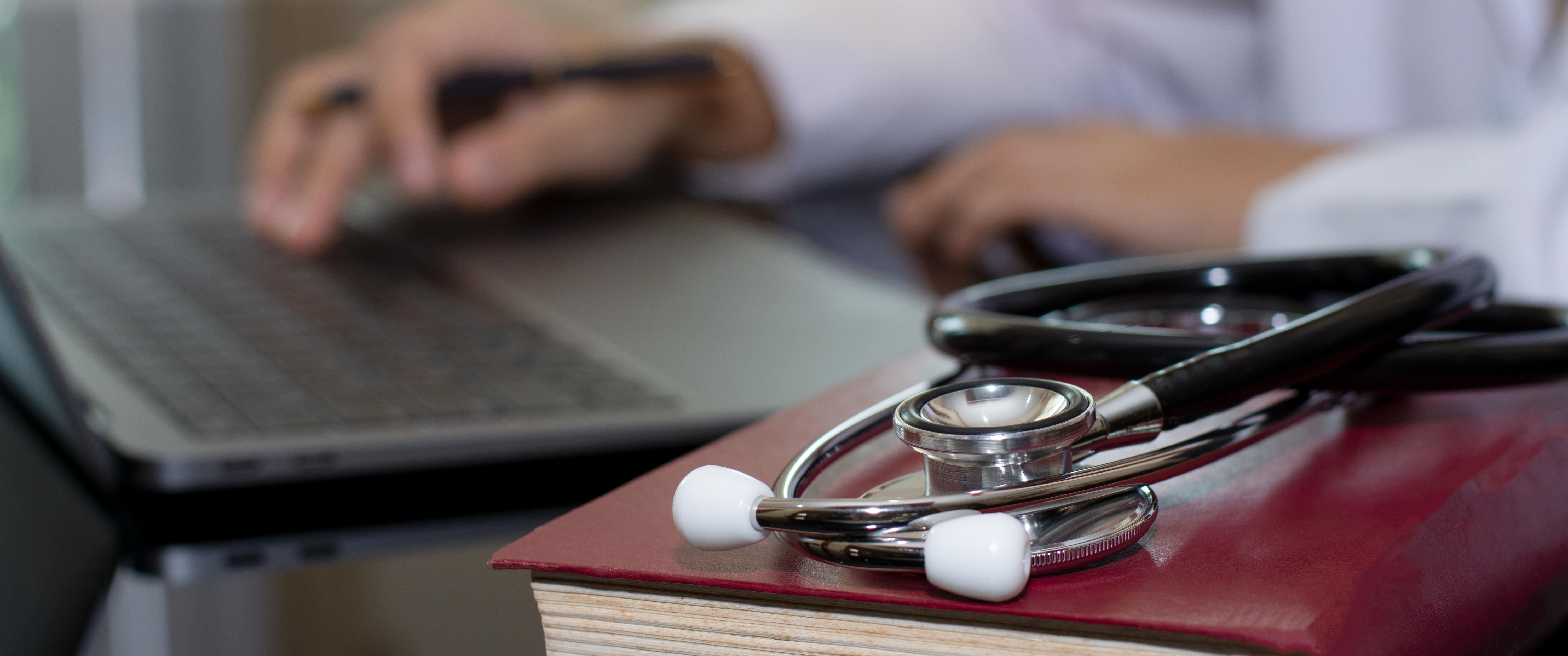 Doctor works on laptop with textbook and stethoscope on the desk 