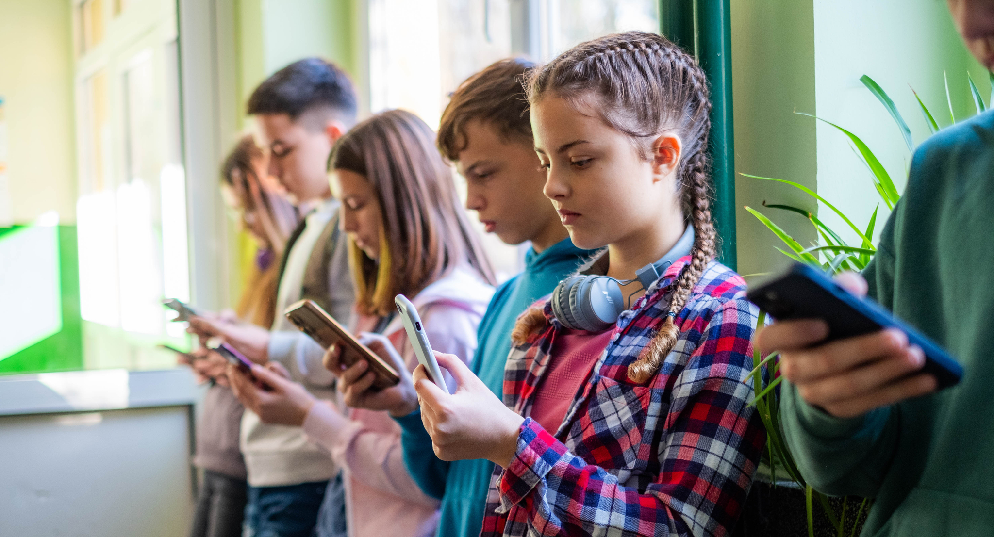 Teenage students are standing in the school hallway, all looking at their phones.