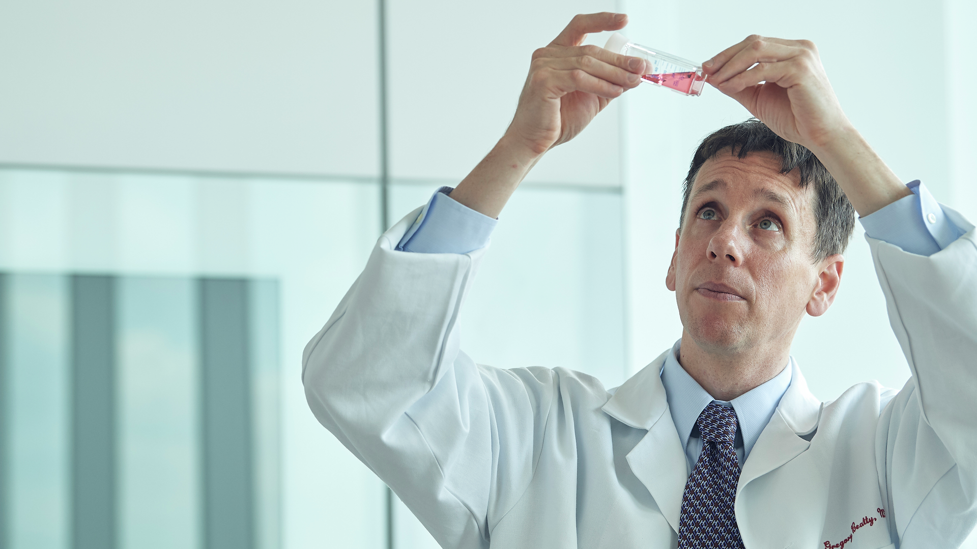 Gregory Beatty in a white lab coat looking up at a vial of liquid