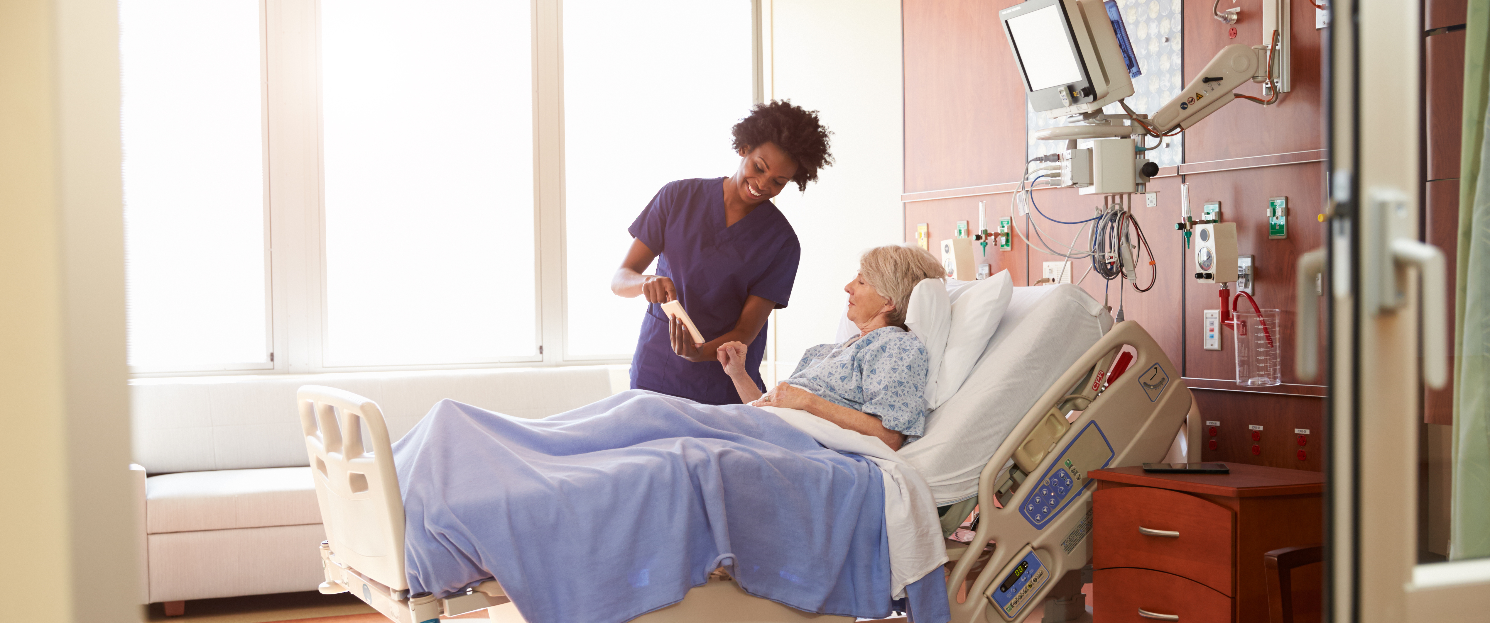 Nurse speaking with patient laying in hospital bed while showing the screen of a tablet.