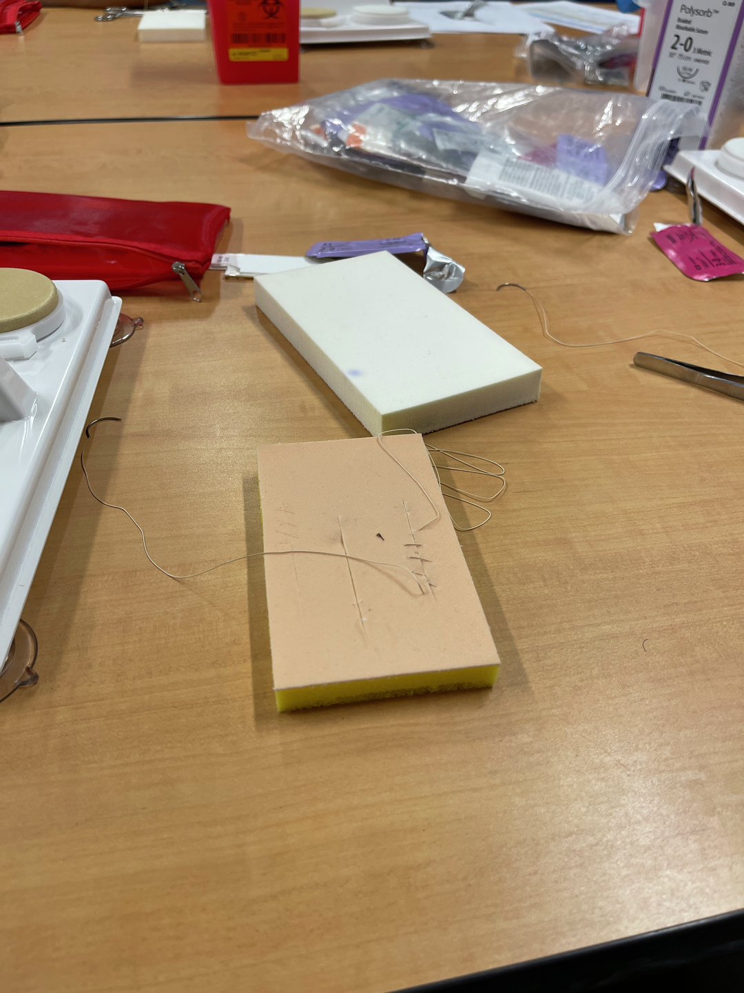 A rectangular piece of foam on a table with thread stitched through it, from someone practicing sewing sutures