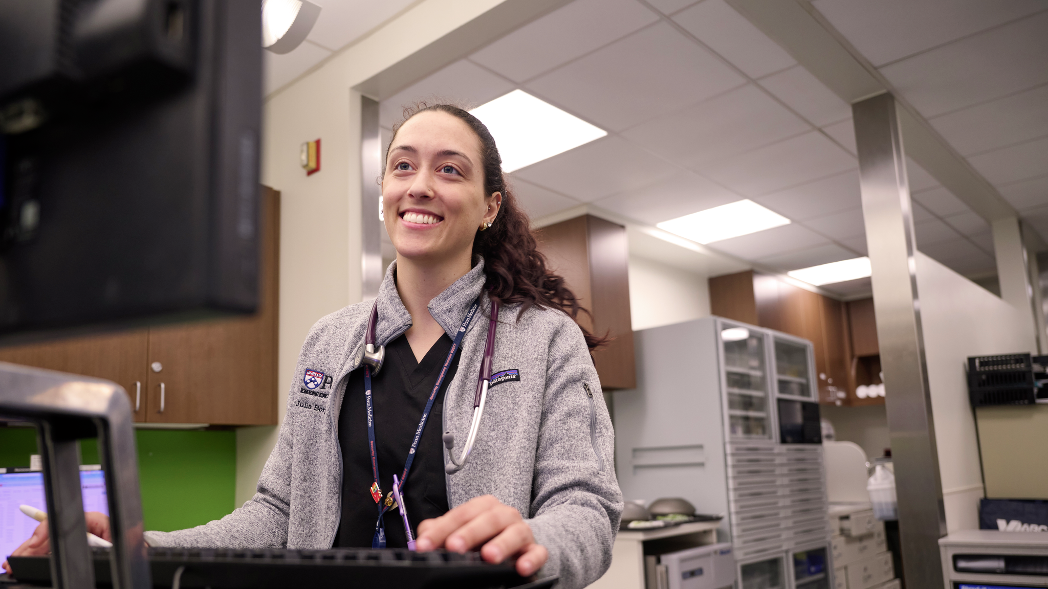 Julia Borgesi smiles while dressed in scrubs looking at a computer monitor