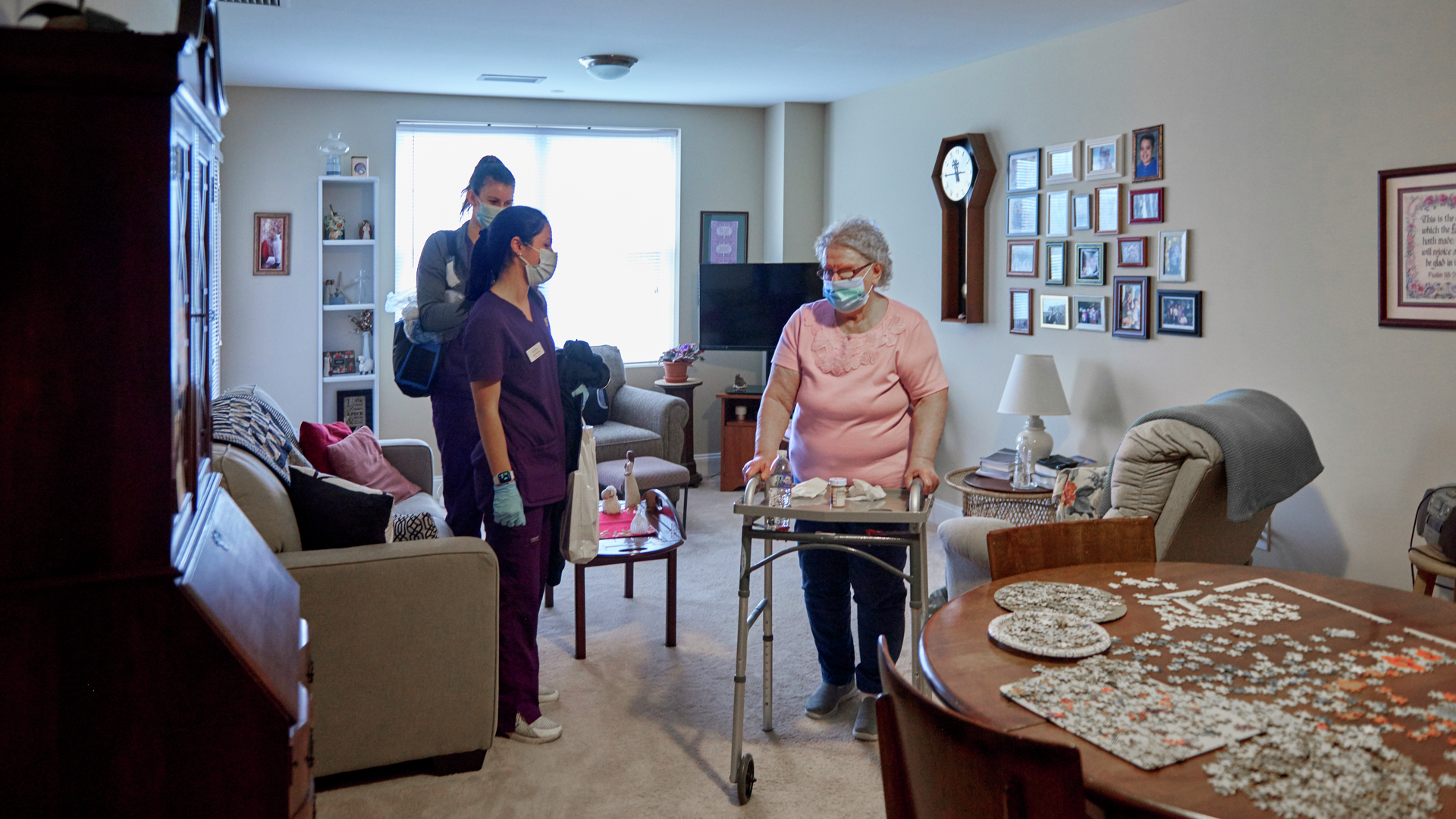 Two nurses stand with an older woman in her living room where she walks with a walker