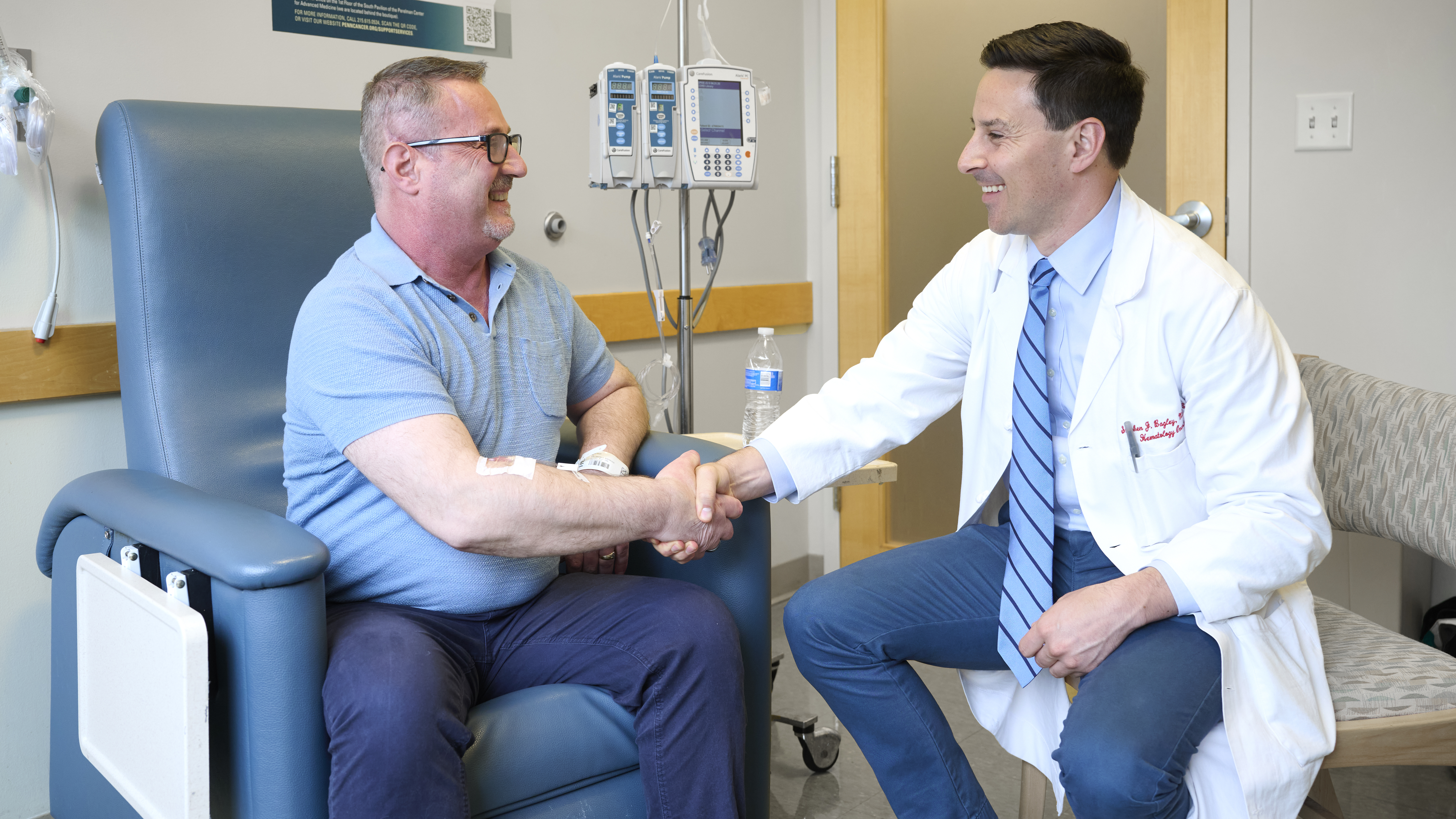 Eduart Cuka and Stephen Bagley, MD, smile and shake hands while seated in an infusion room