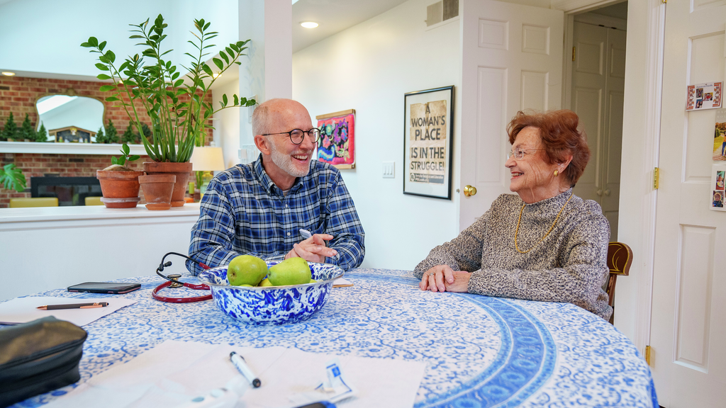 Leon Kraybill, a smiling man with a white beard, sits at a kitchen table talking with an older woman patient with stethoscope and other medical items visible.