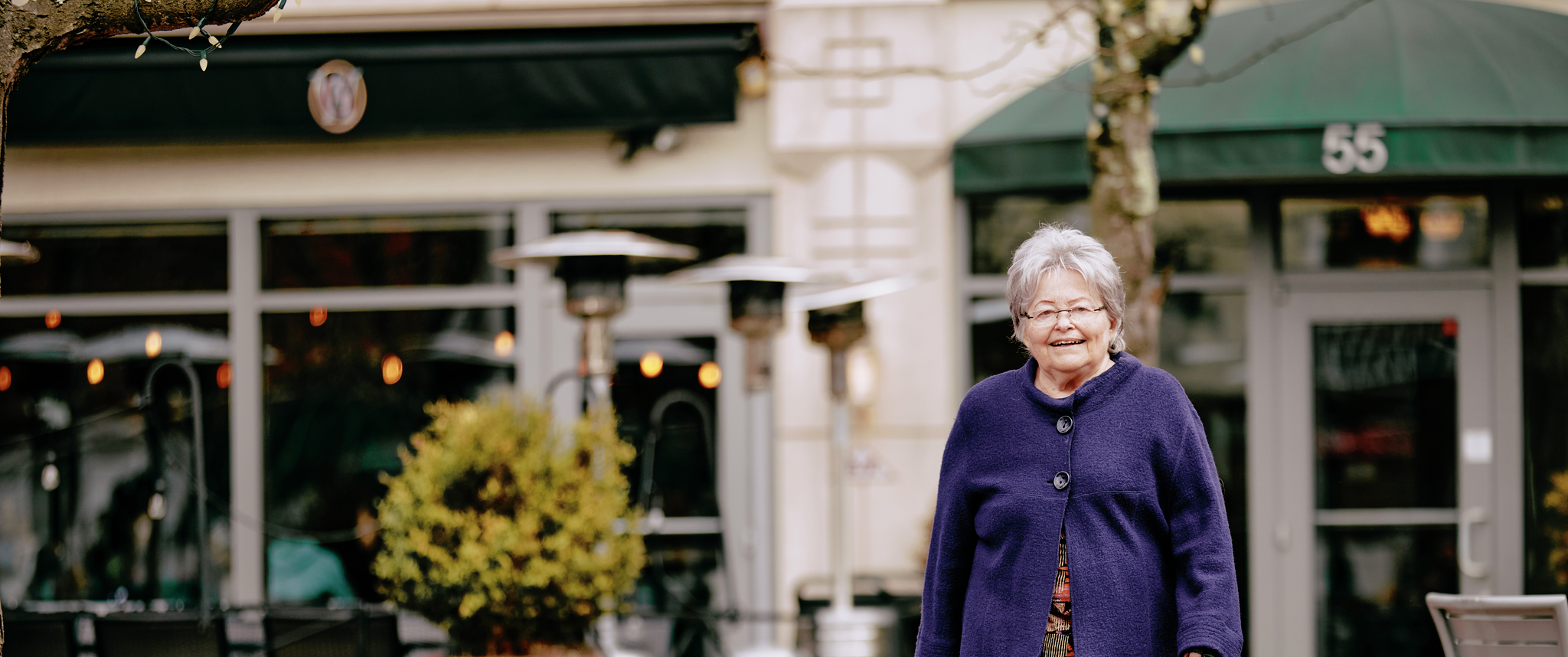 Mimi Reed, a senior woman wearing a purple cardigan, smiles while walking in a downtown setting