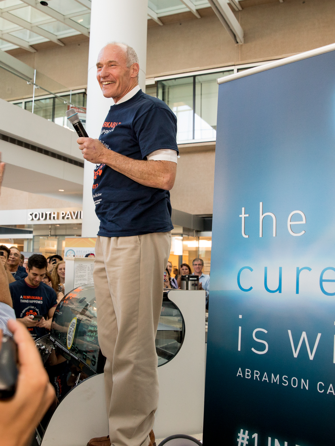 Carl June, MD, wearing an "A Remarkable Thing Happened" T-shirt, stands holding a microphone in front of a flash mob crowd celebrating his discovery being approved by the FDA