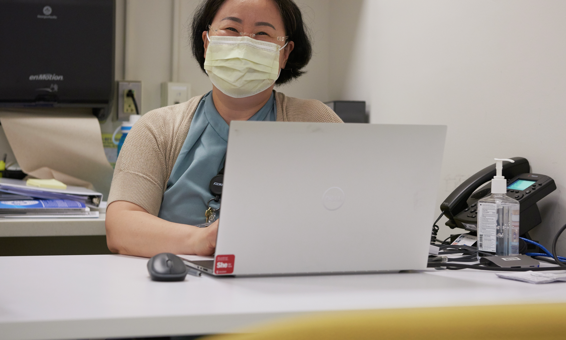 Helen Jeong smiles while masked and seated behind a laptop