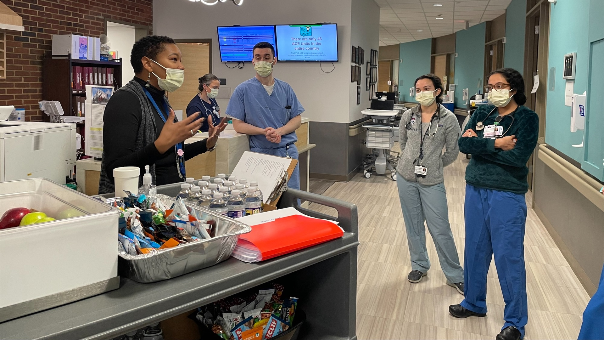 Kenya Pitt stands next to a cart filled with water bottles and healthy snacks, talking with a small group of employees in scrubs in a hospital hallway