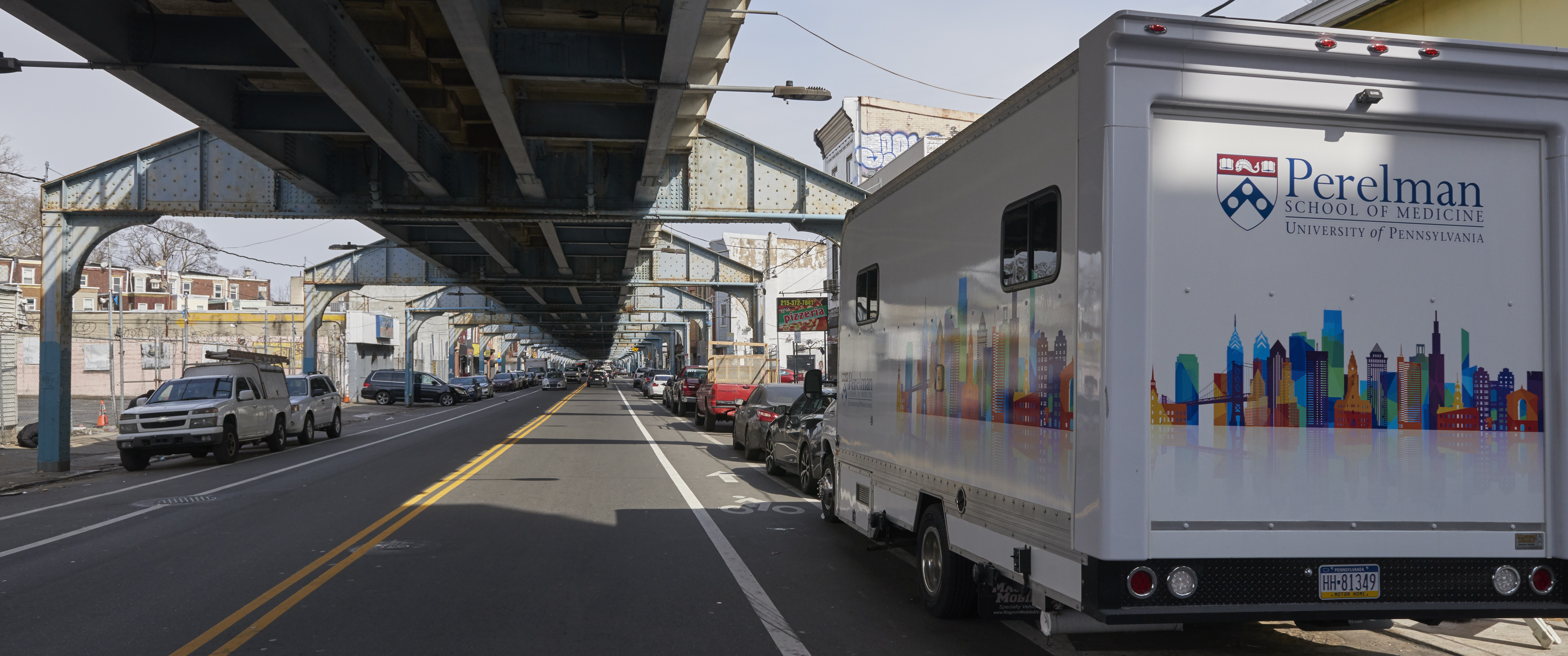 A large truck with a Penn logo and colorful city skyscape graphic is parked in the shadow of elevated train tracks on Kensington Ave