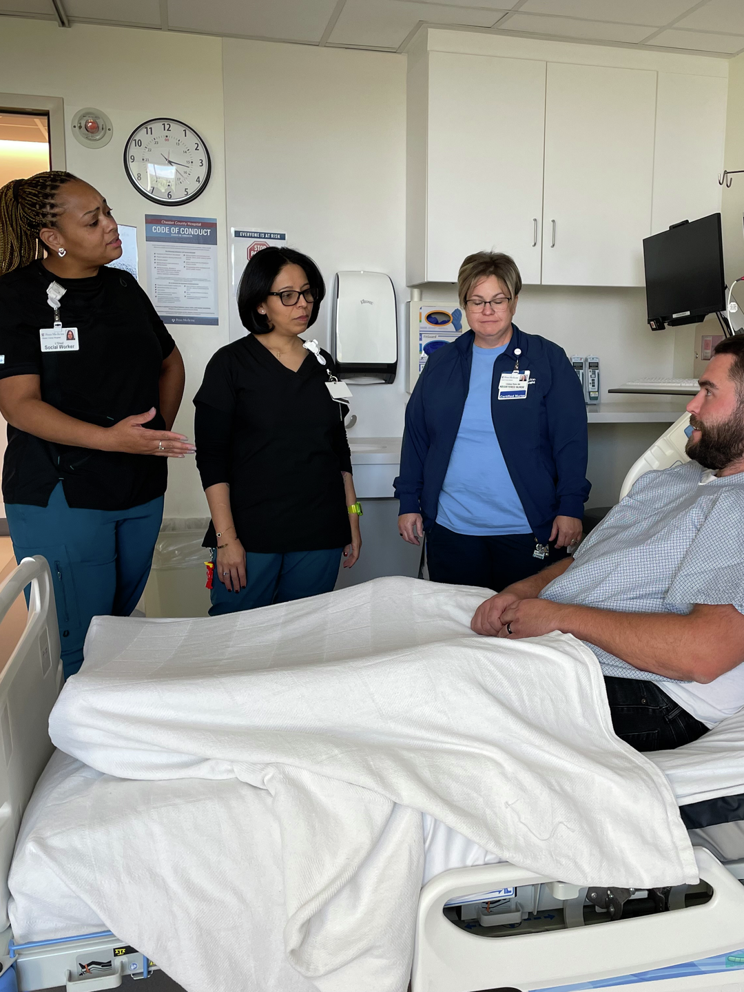 Three team members stand beside a patient in a hospital bed