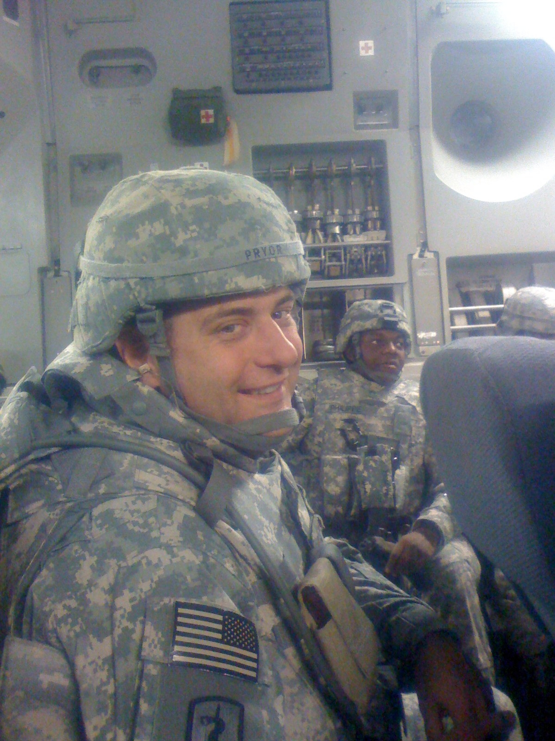 John P. Pryor, MD, dressed in camouflage gear and helmet, smiles at the camera with another military member in the background.