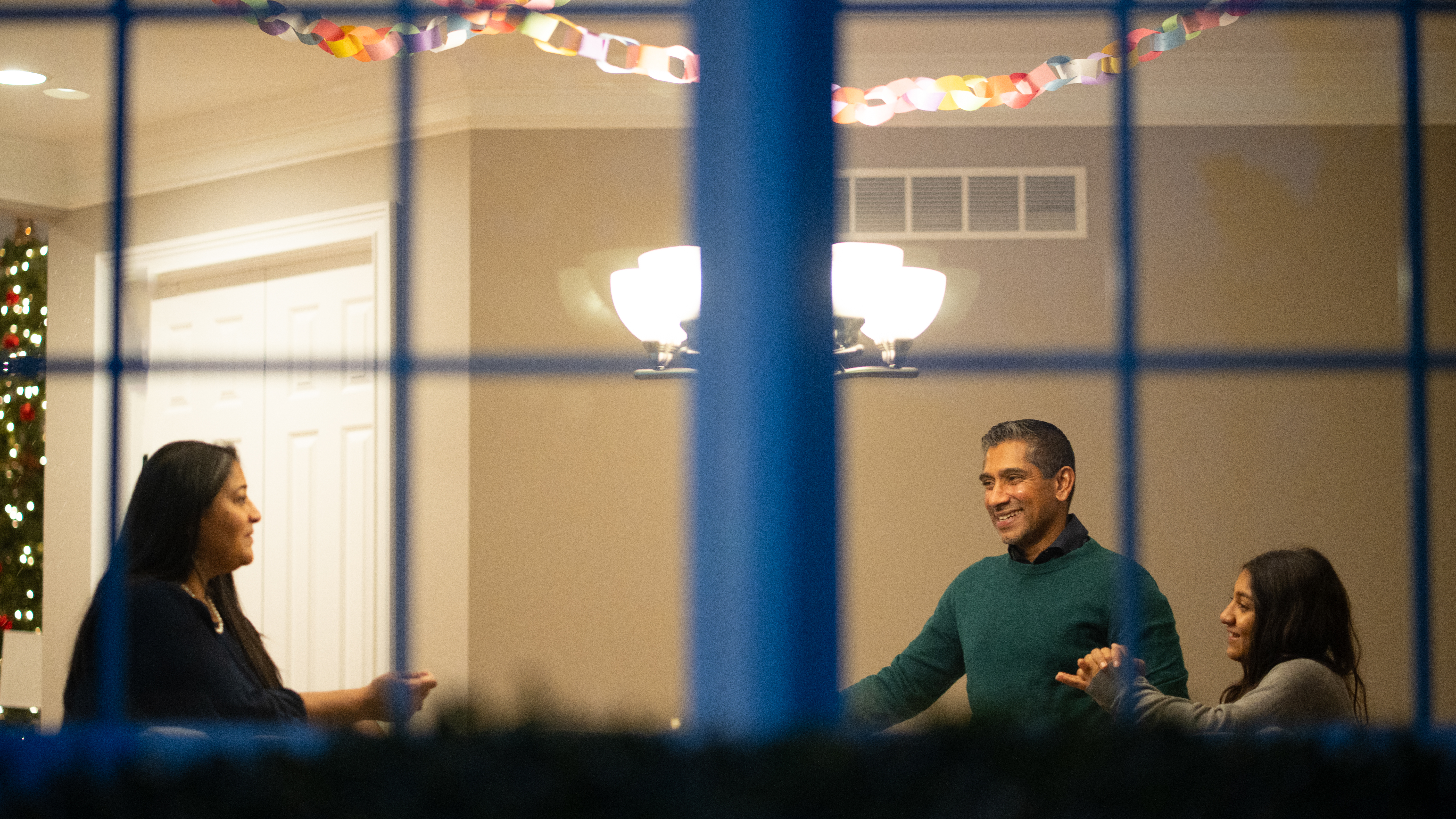 Aileen John, her husband, and daughter are talking together, seen through an exterior window of their home