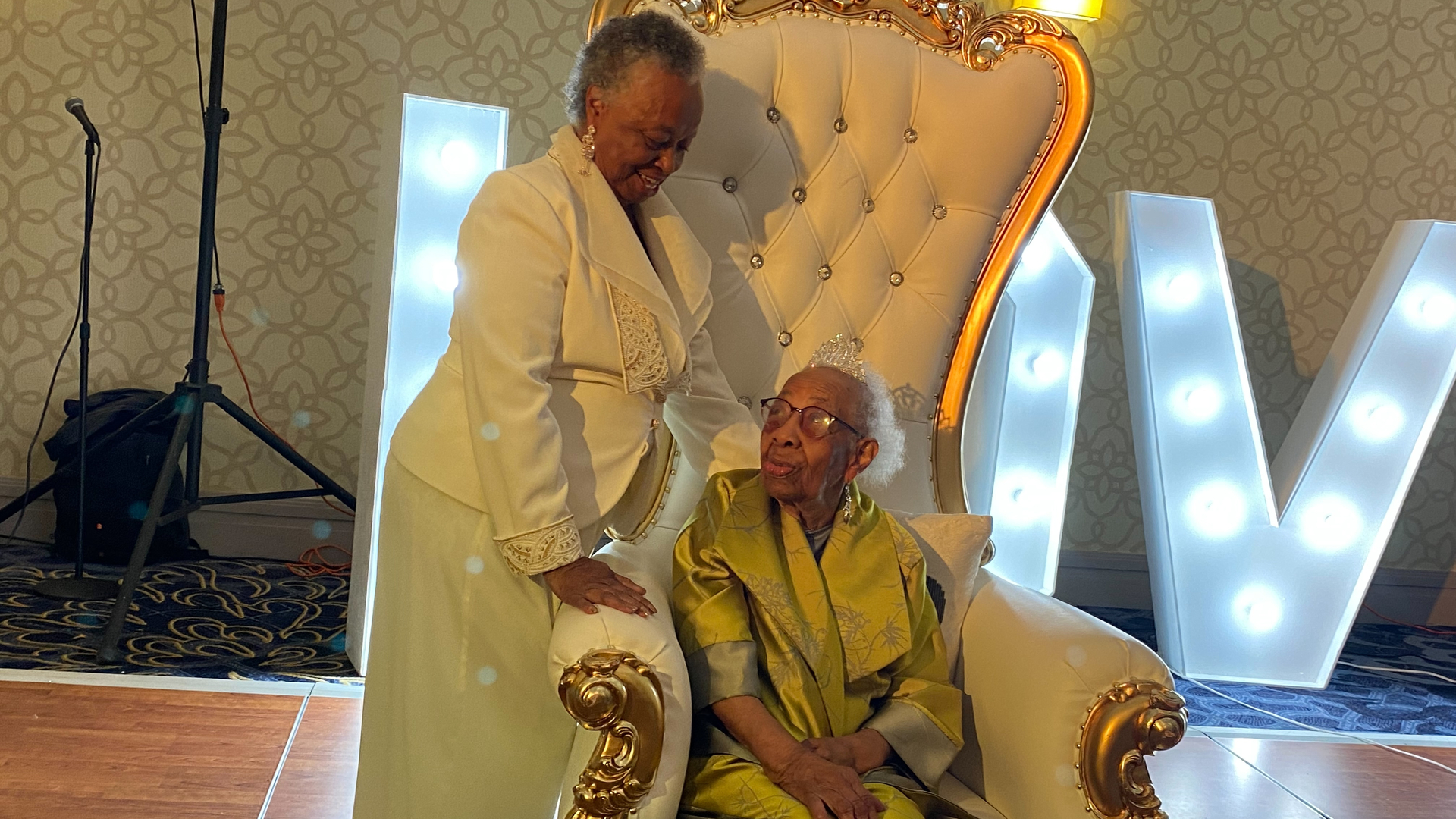 100-year-old Eloise Brown, wears a tiara and a silk kimono dress sitting in an oversized plush throne, with her daughter Joan Reid standing beside her in a white pantsuit.