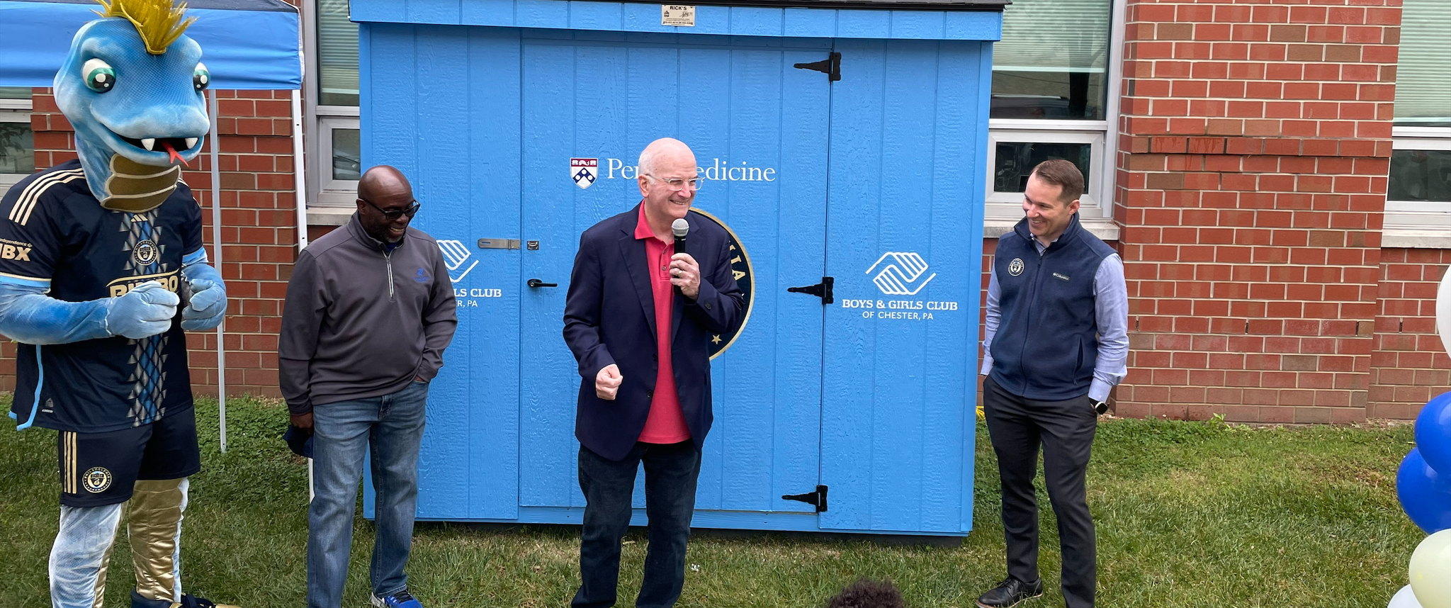 Kevin Mahoney holds a microphone standing in front of a bright blue shed (community food pantry), surrounded by Philadelphia Union officials and mascot Phang