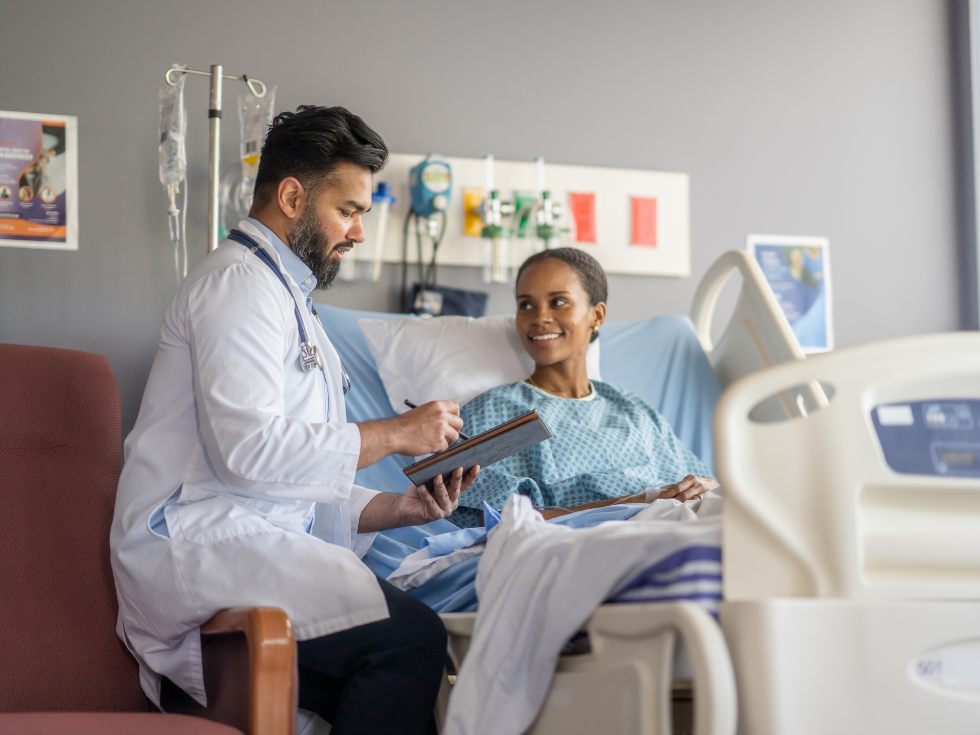 A woman sits up in a hospital bed wearing a gown as she talks with her male doctor about her surgery