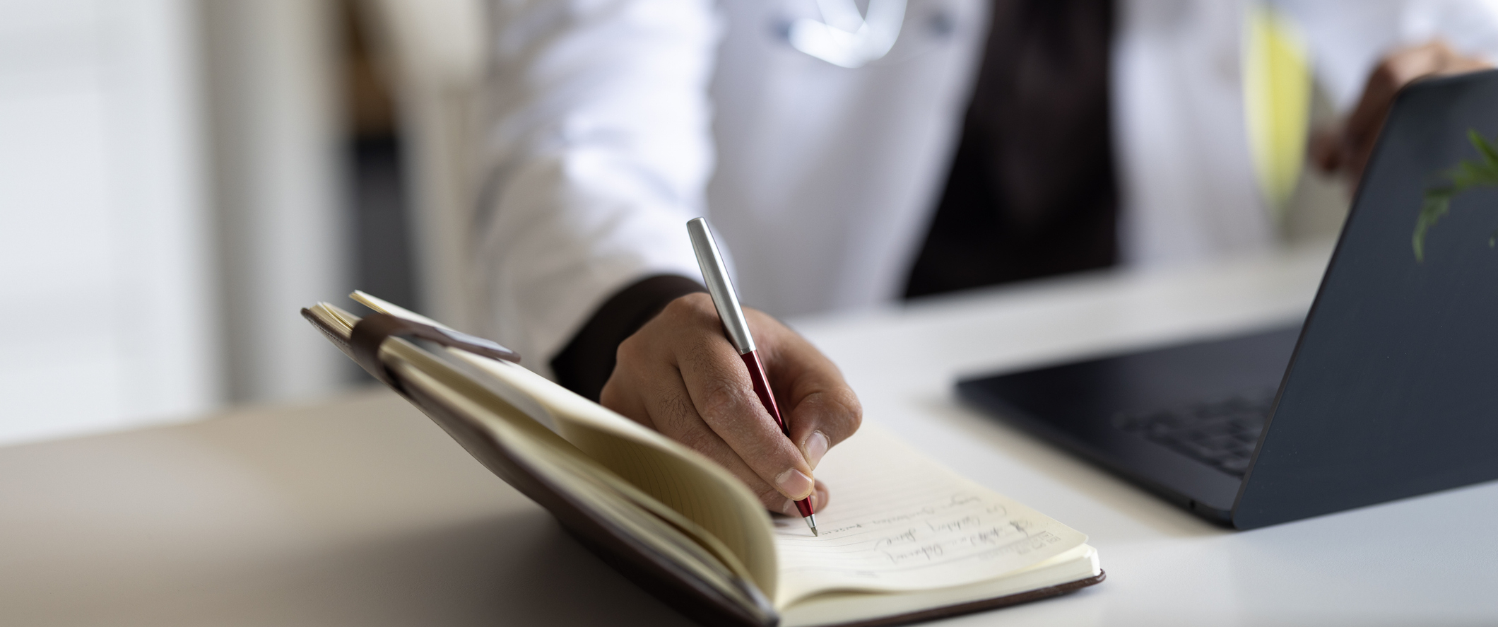 A doctor sitting in front of a laptop and writing in a journal