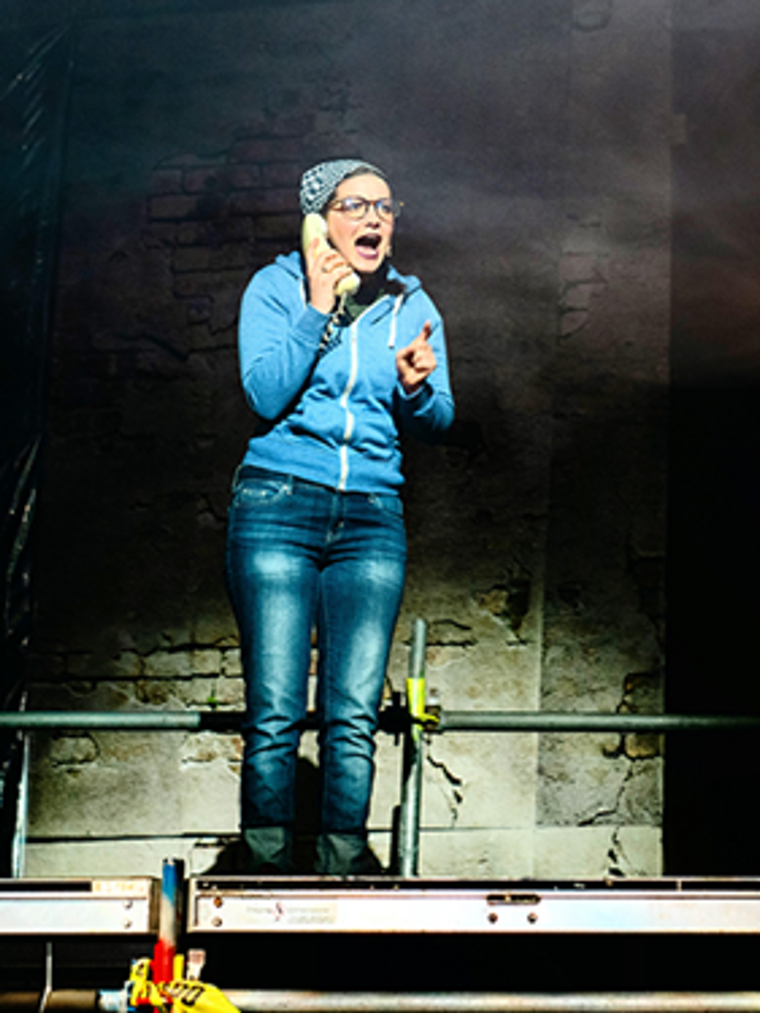 Hannah Parke on stage, wearing a blue hoodie and holding a telephone, against a dark background.