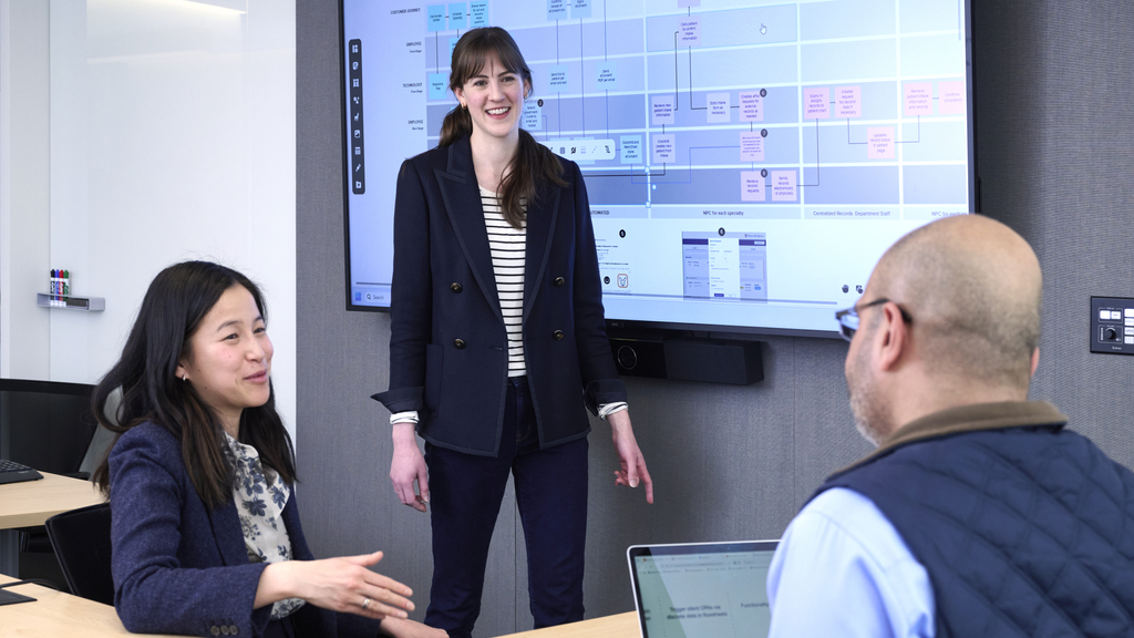 Lauren Hahn stands in front of a large screen in a conference room with Kat Lee and Mohan Balachandran 