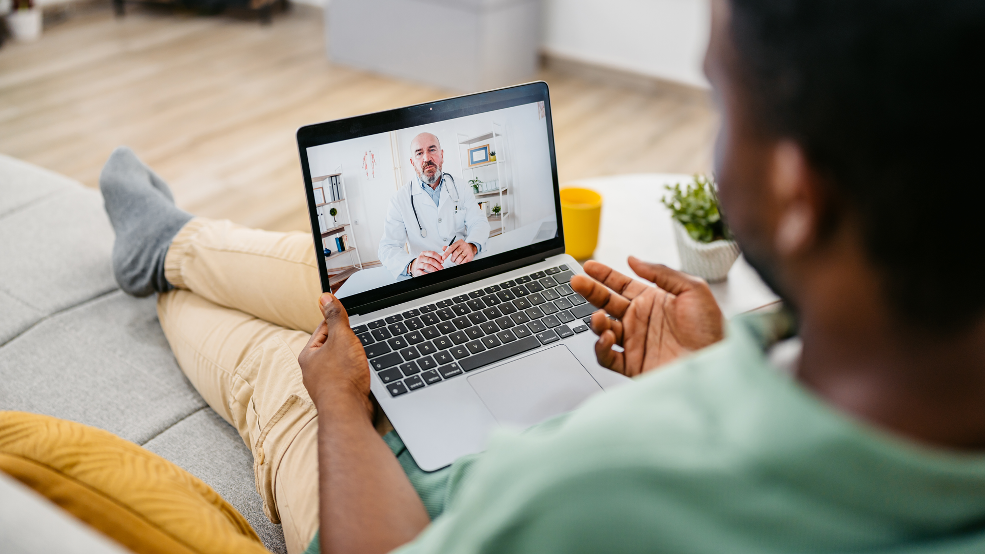 Young black man sitting on the sofa in the living room and having a doctor's appointment online via laptop
