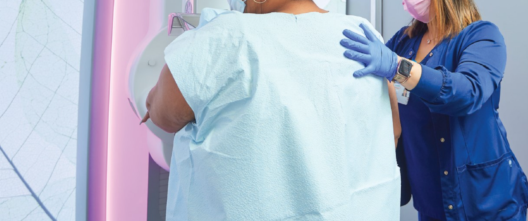 A woman stands with her back to the viewer, her front in a mammography machine, while a technician places a hand on her shoulder