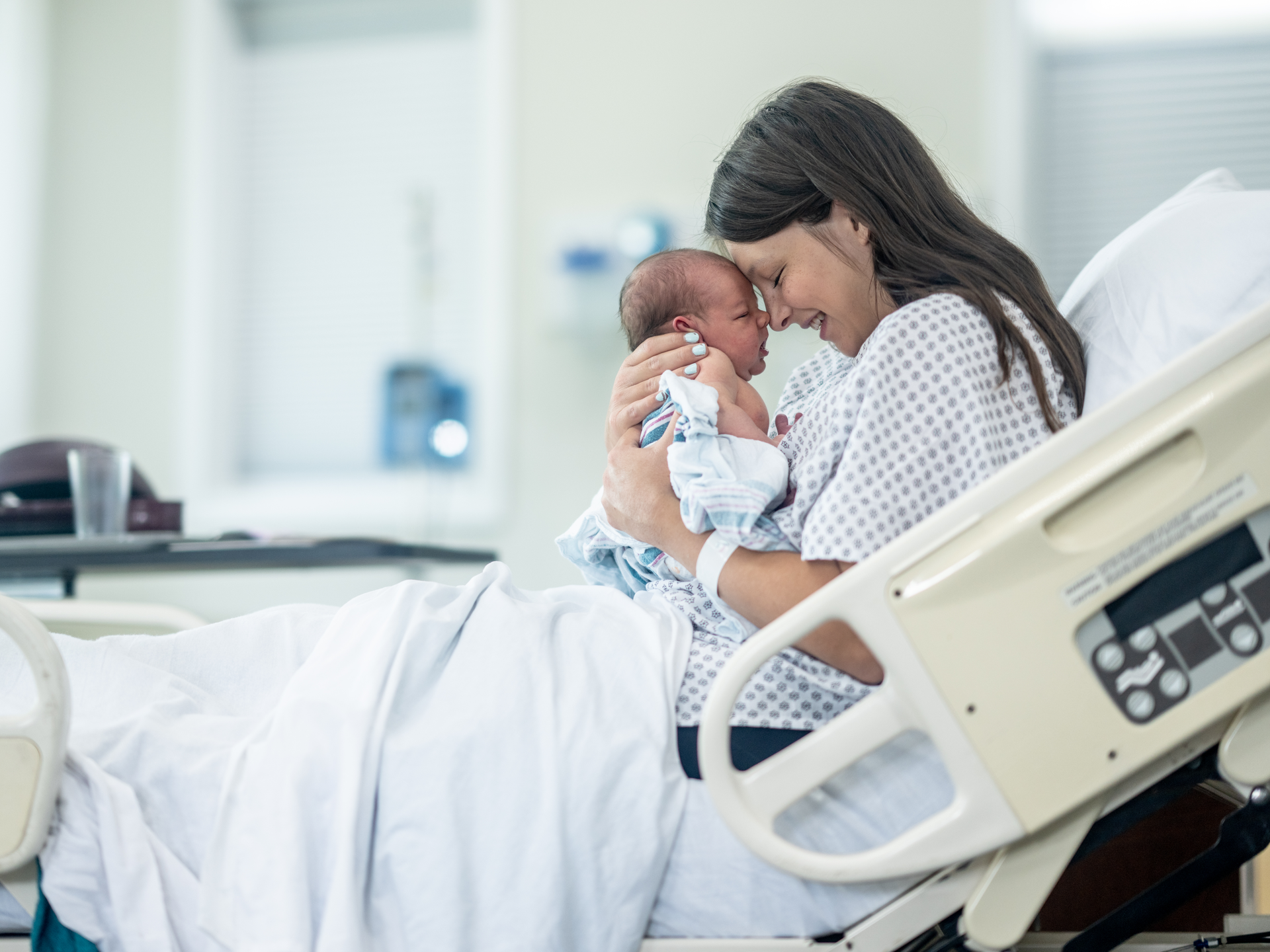 A new mother sits up in hospital bed shortly after delivery as she holds her newborn out in front of her 