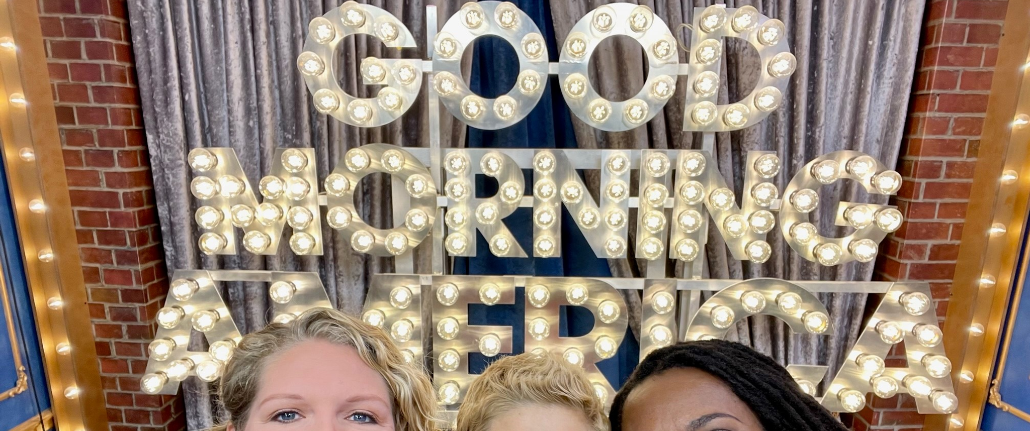 Three women pose for a group selfie photo in front of the Good Morning America Logo made of lights: Hayley Knollman, Kate Korson, and Lola Fayanju