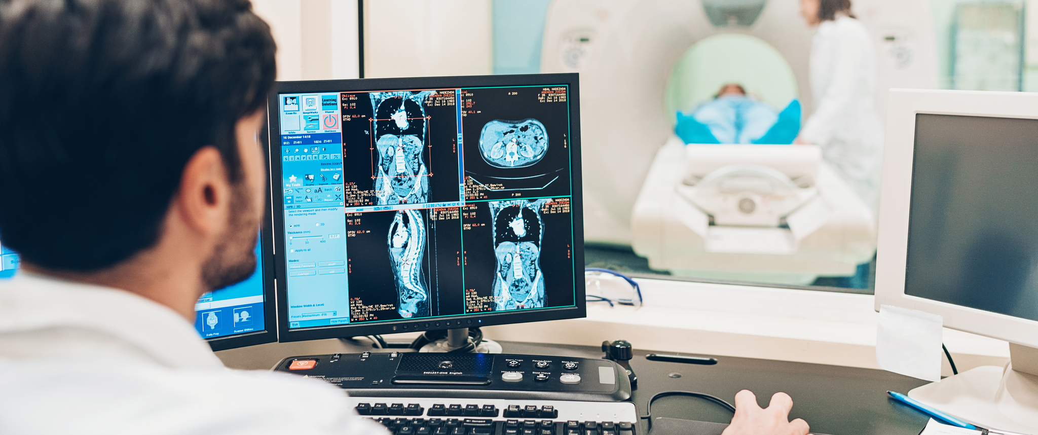 The back of a doctor looking at scans of an abdomen on his monitor while a patient recieves a CT scan in the background.
