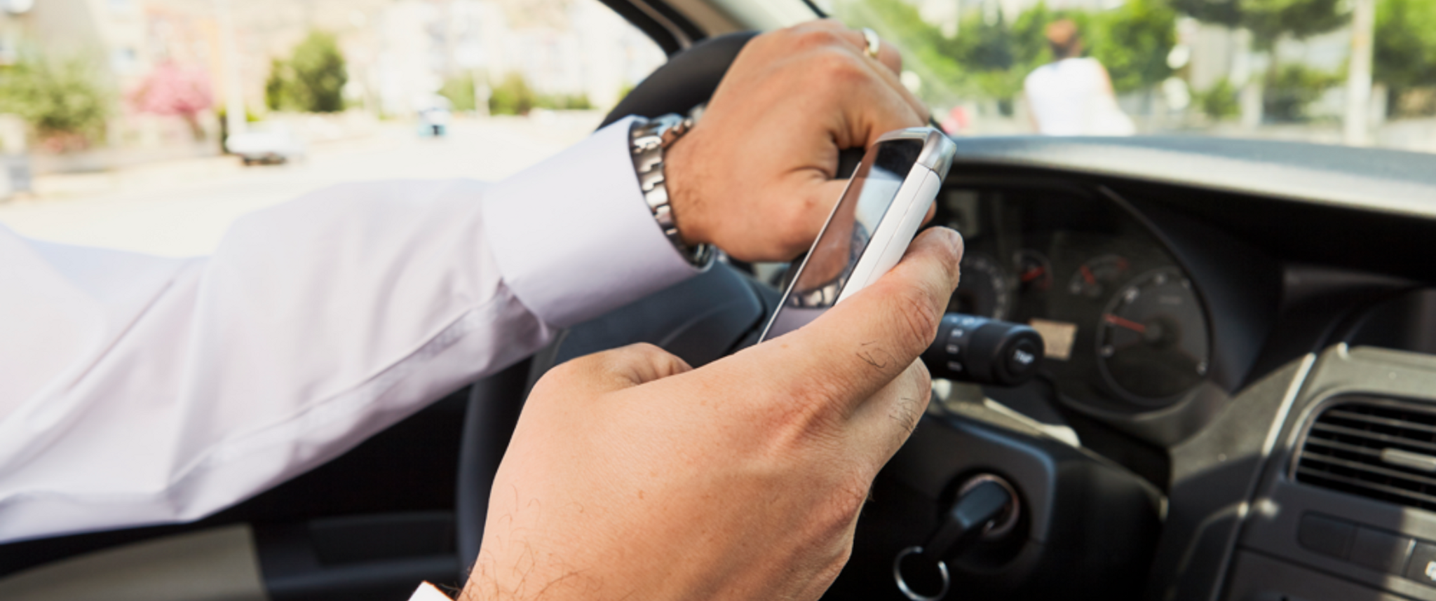 A person's left hand holds a steering wheel in a car while the right hand holds a cell phone
