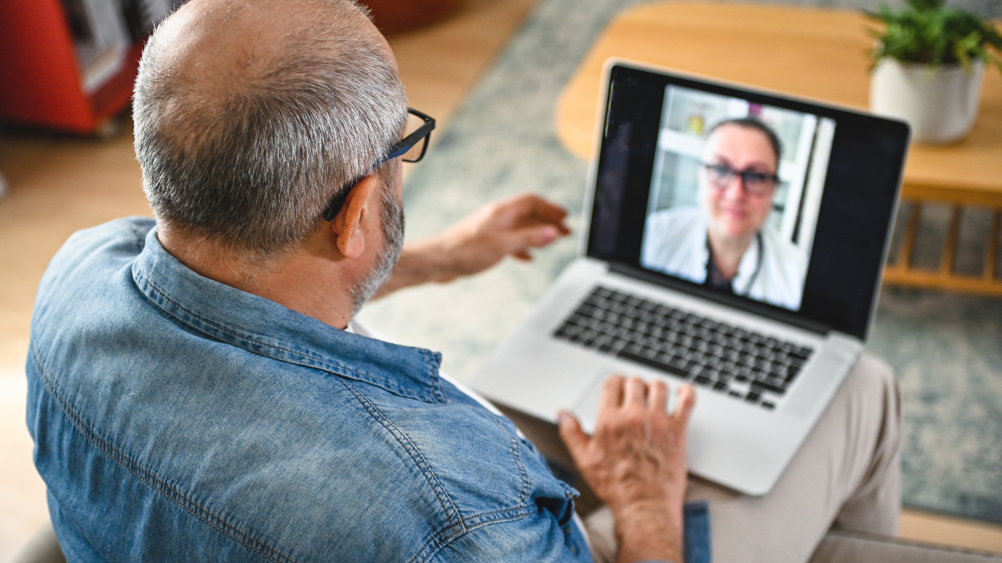 An adult man sits in the living room and communicates with a doctor via video link on a laptop.