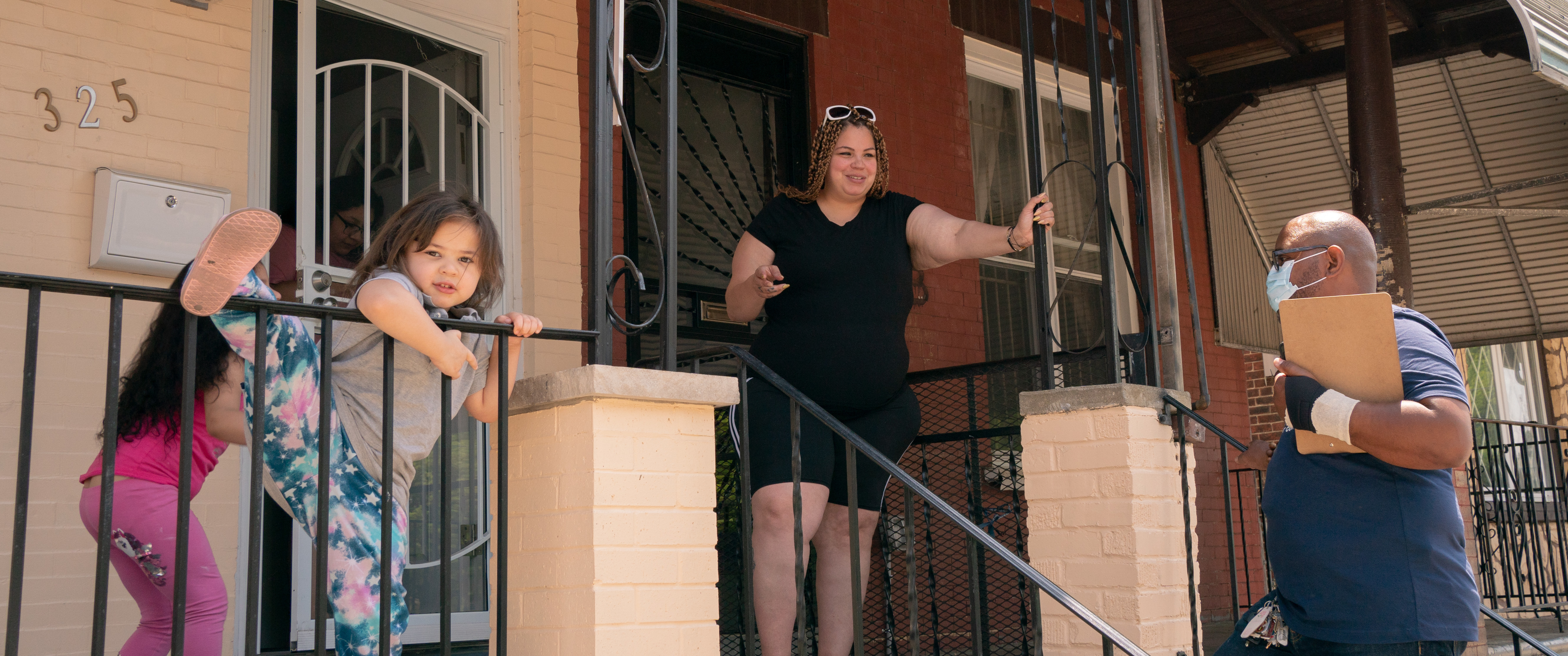 Two young girls climb and jump on a porch while Cristal LaTorre stands at her front door speaking with Yuhnis Sydnor, on the front steps