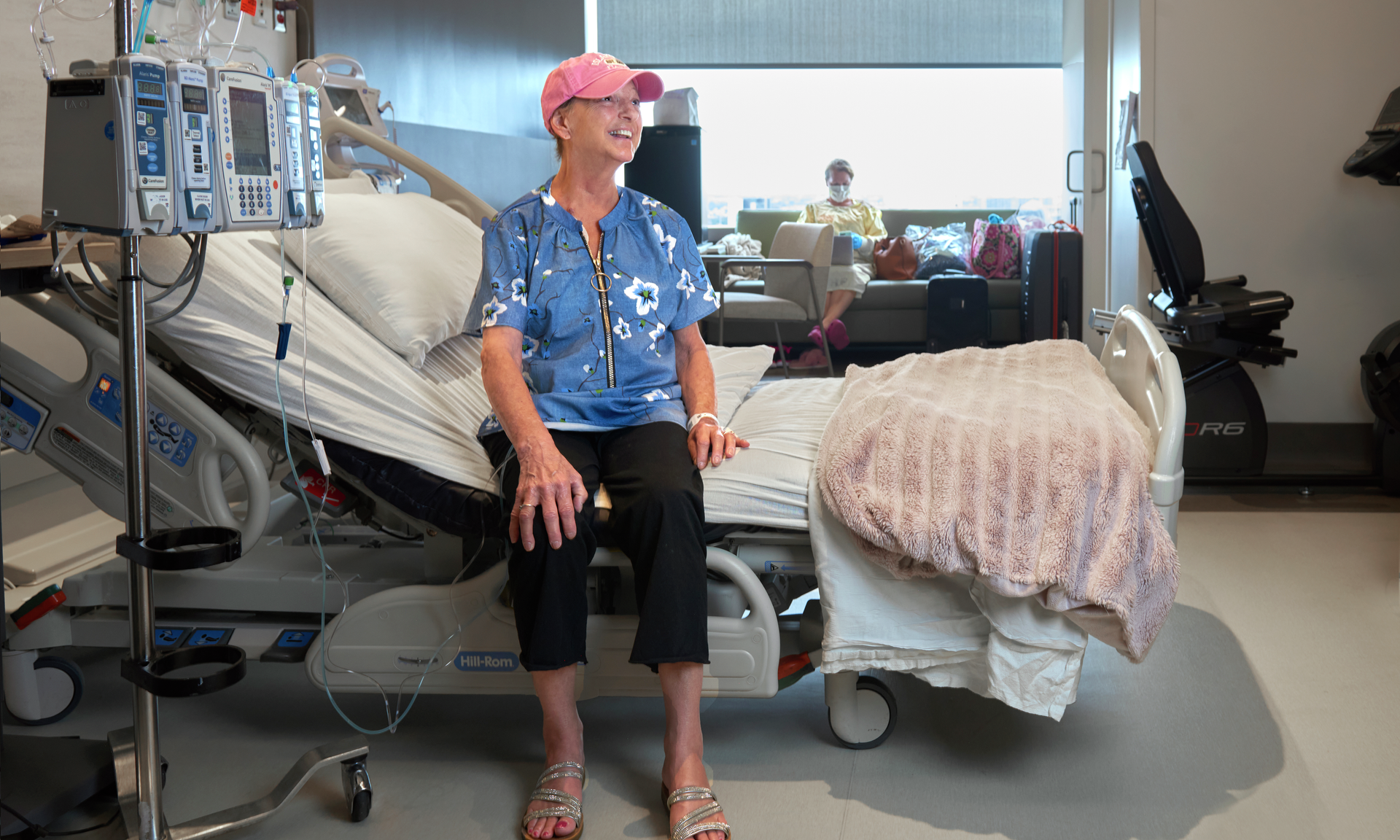 Peggylee Giles, smiling wearing a pink cap and sparkly sandals, sits on her hospital bed. Daughter Jessica Swal in a yellow gown types on a laptop behind her