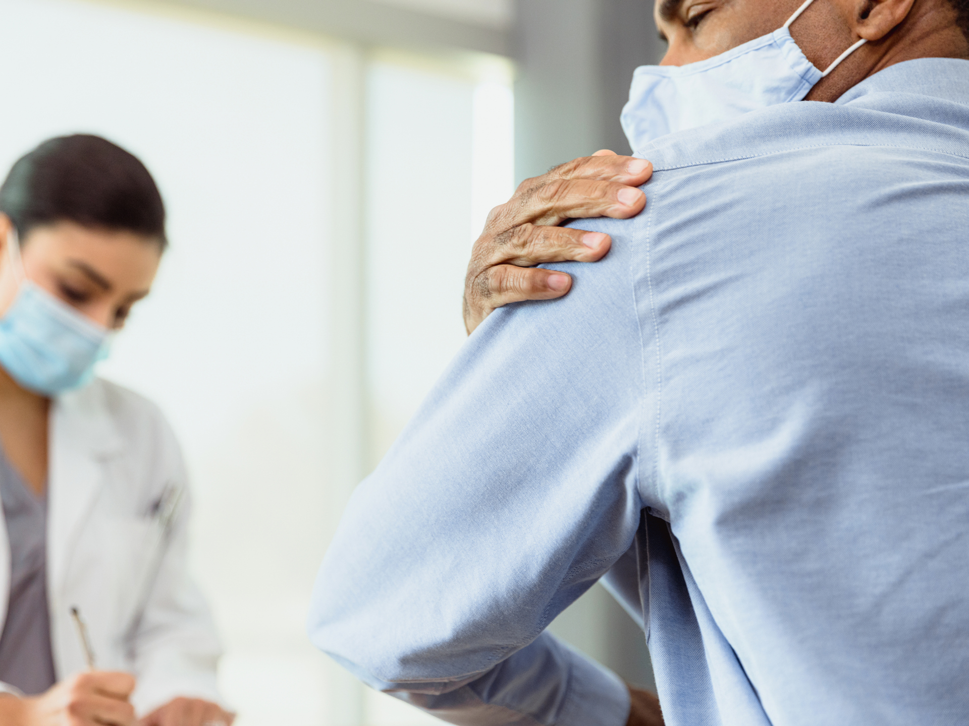 A mature man, experiencing shoulder pain, rubs his shoulder while talking with a female doctor. The doctor is taking notes in the background.