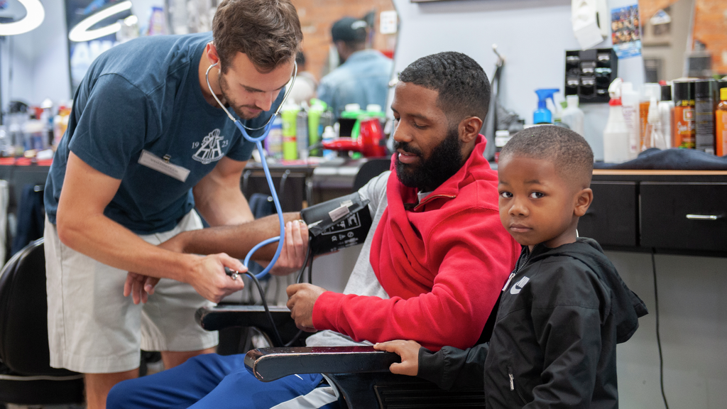 A medical student measures a young Black man's blood pressure while the man's approximately 4-year-old son stands nearby