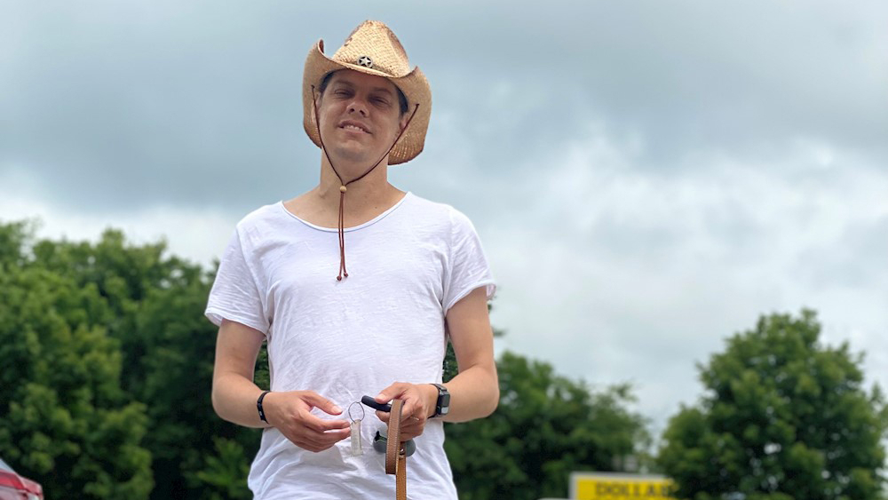 Donald Philp stands facing the camera wearing a cowboy hat, with trees and sky filling the background