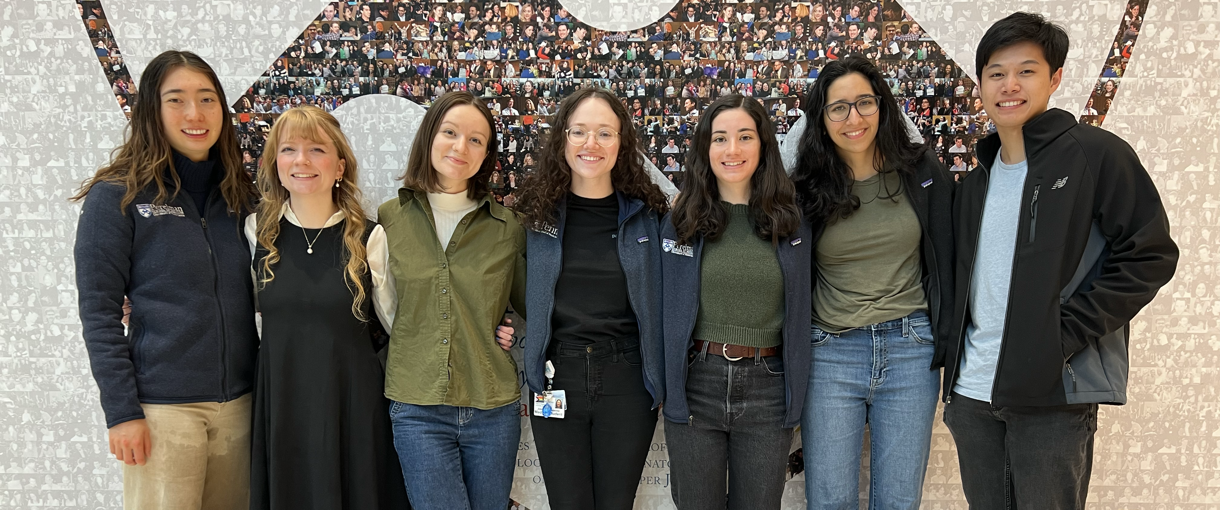 A group photo of medical students Catherine Shen, Elizabeth Whidden, Phoebe Cunningham, Jess Campanile, Sarah Wornow, Isha Thapar, and Jonathan Szeto standing in front of a photo collage in the shape of the Penn shield.