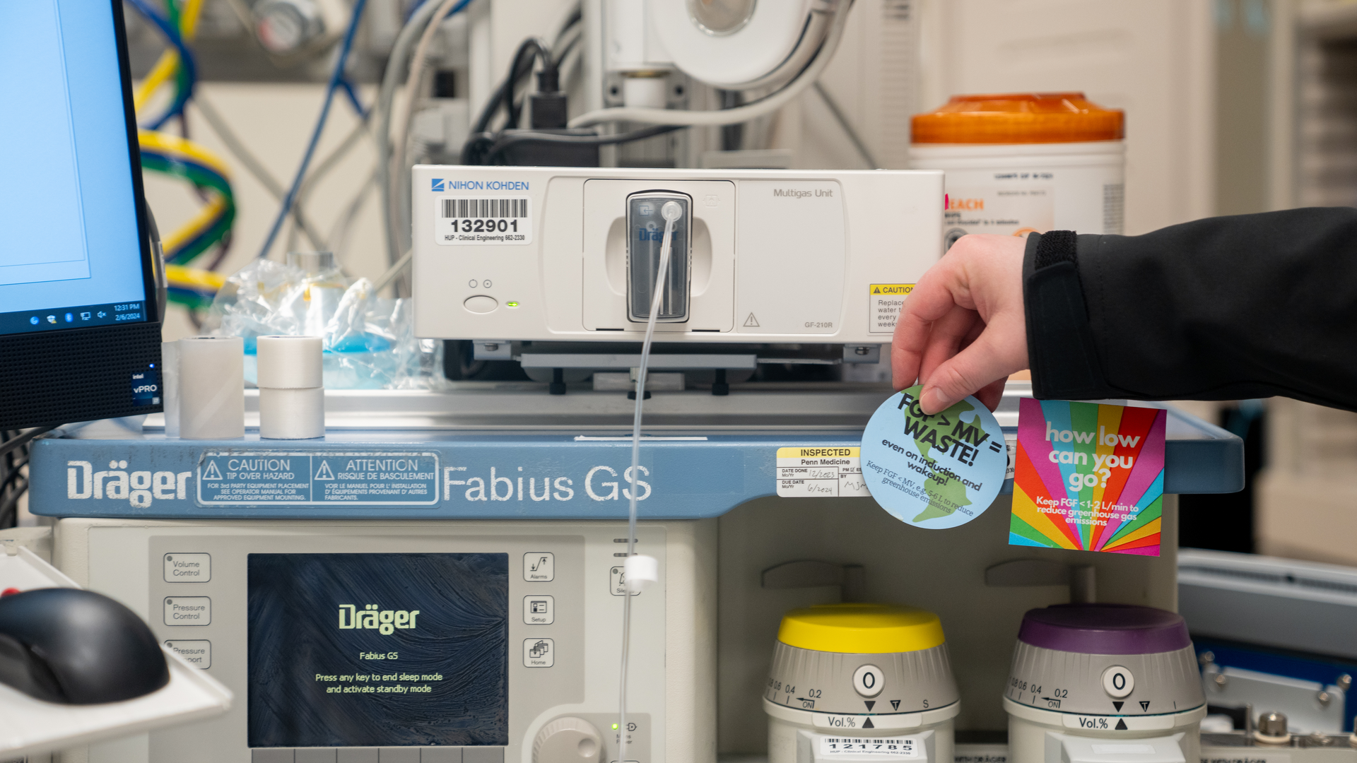 A hand holds up a round magnet with an Earth design and text saying 'FGF>MV=WASTE' to encourage anesthesiologists to adjust flow settings.