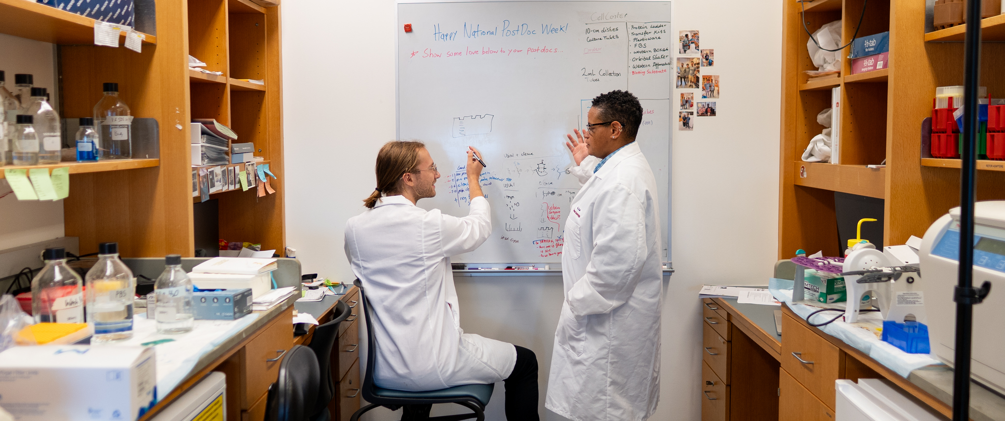 Donita Brady speaking with a colleague who is writing on a whiteboard in a lab.