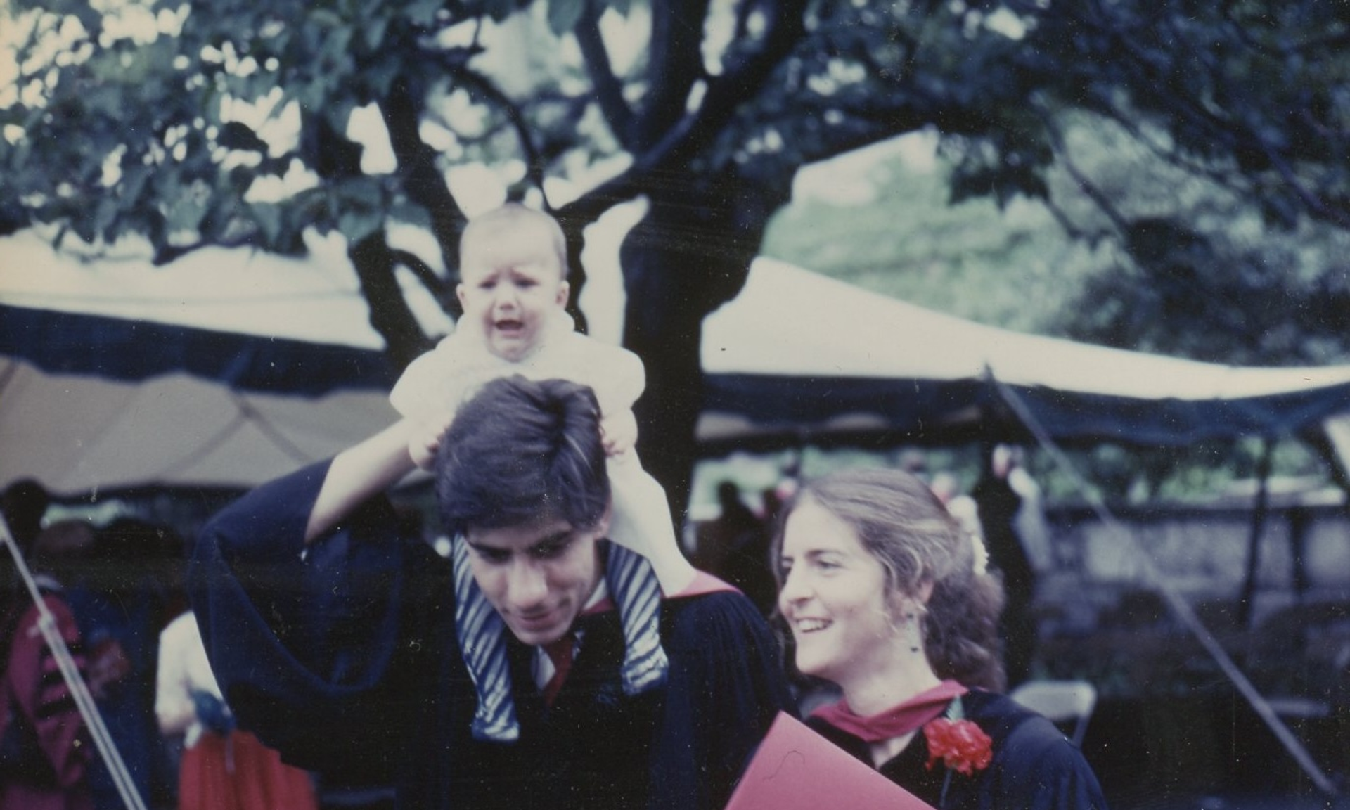 A young Al Maguire holds a baby atop his shoulders, standing next to a young Jean Bennett, both in graduation regalia