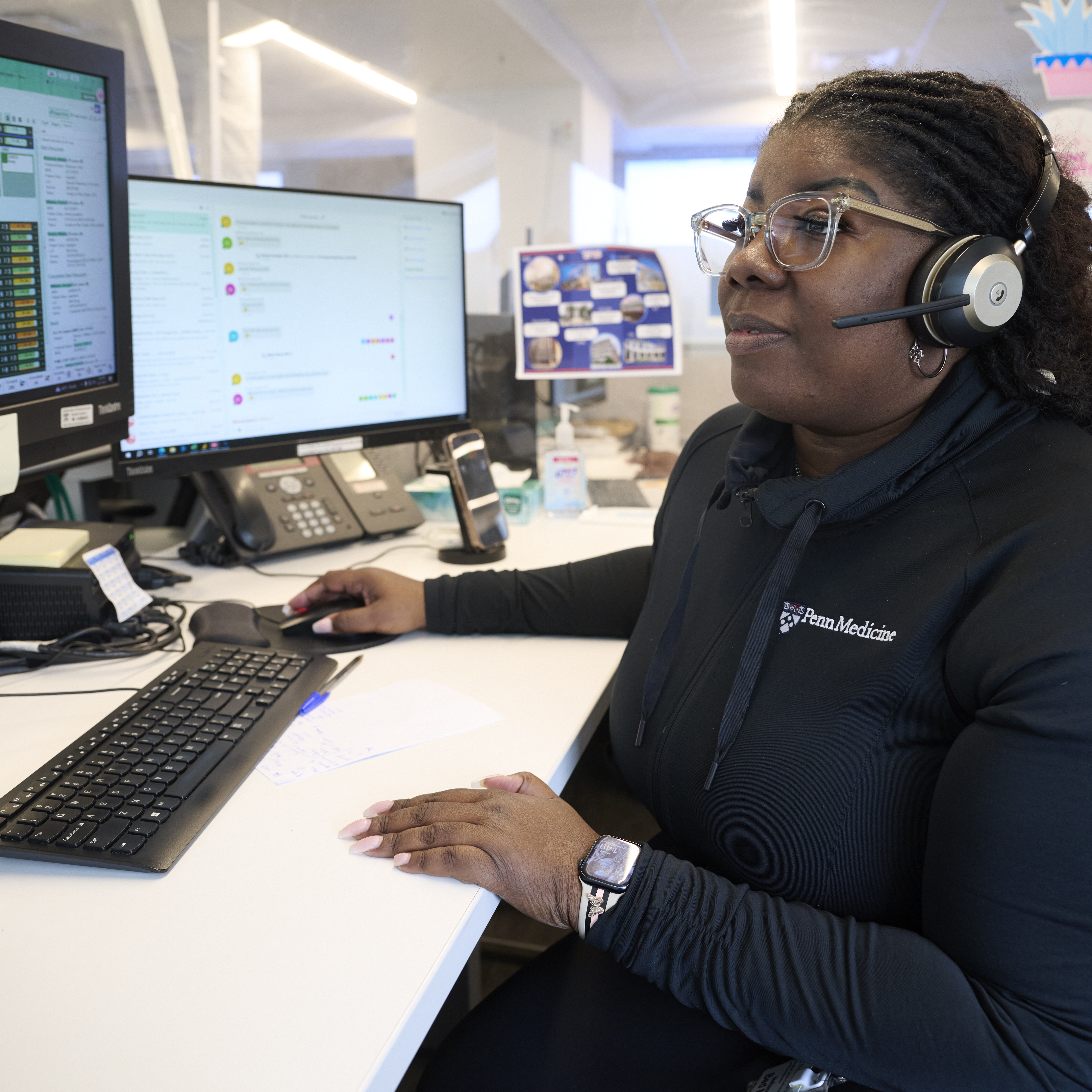 A woman wearing a headset, glasses, and a Penn Medicine jacket sits at a computer desk