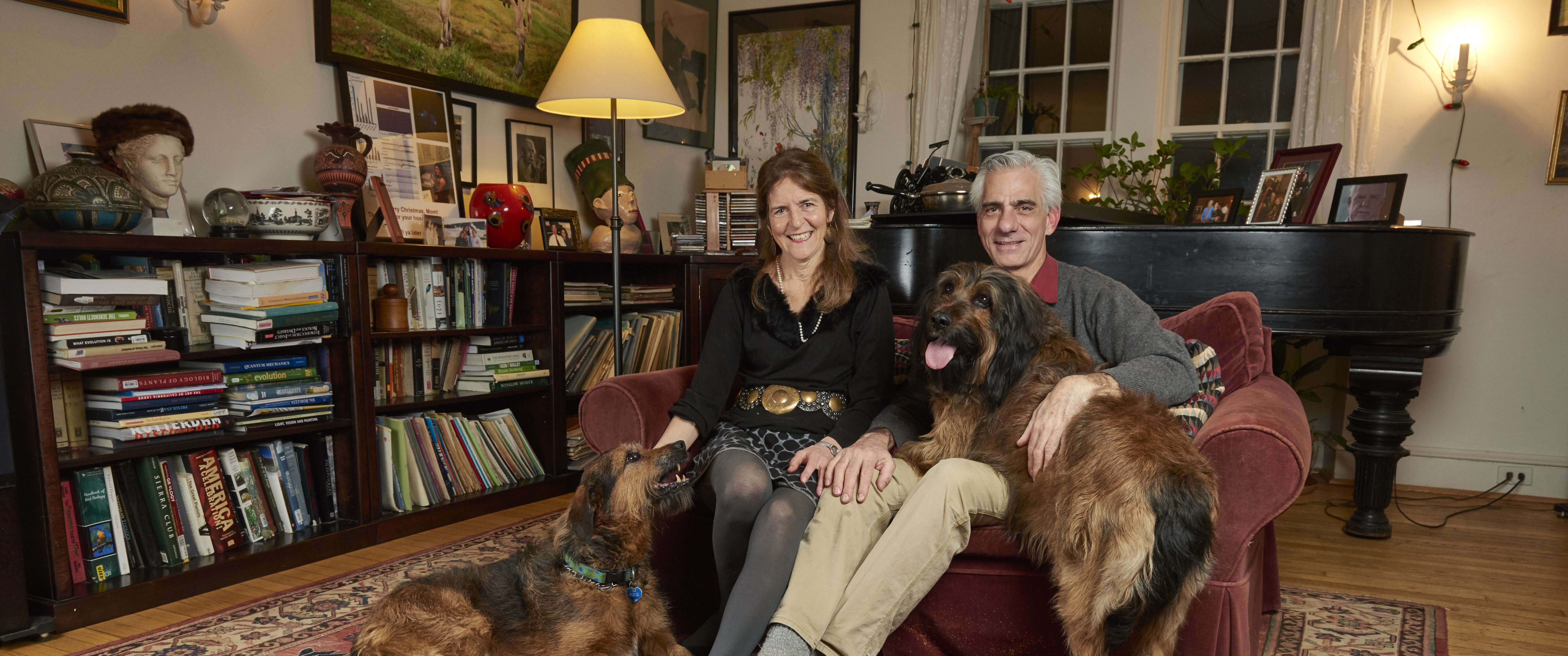 Jean Bennett and Albert Maguire sit on their couch with one dog at Bennett's feet looking up and another large dog on Maguire's lap