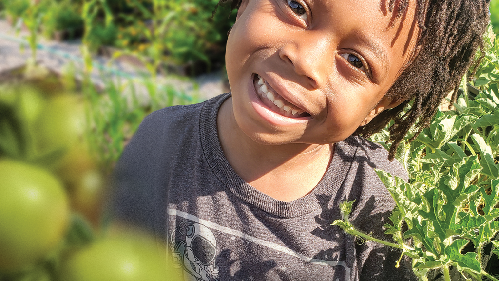 A little boy smiles, surrounded by green growing produce in a garden 